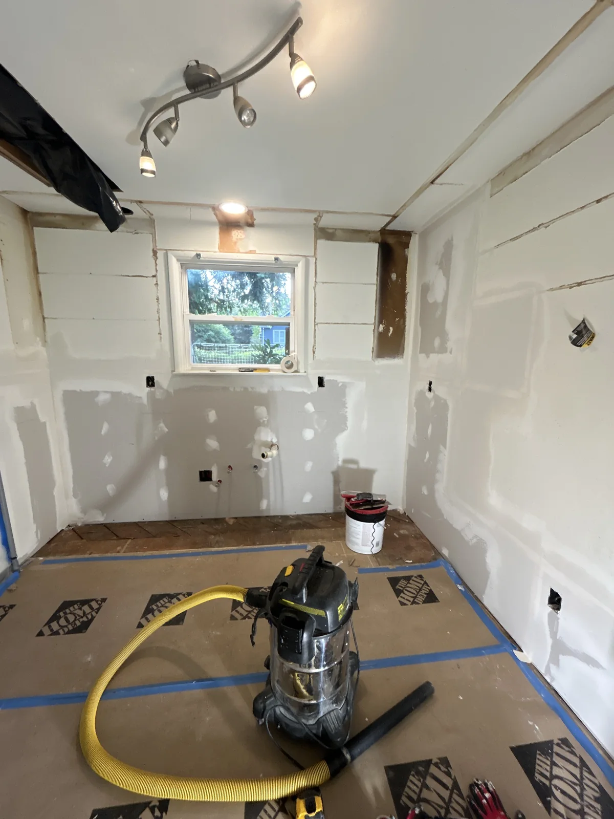 A shop vacuum with yellow hose rests on paper-covered floor in a room with patched drywall walls and a window.