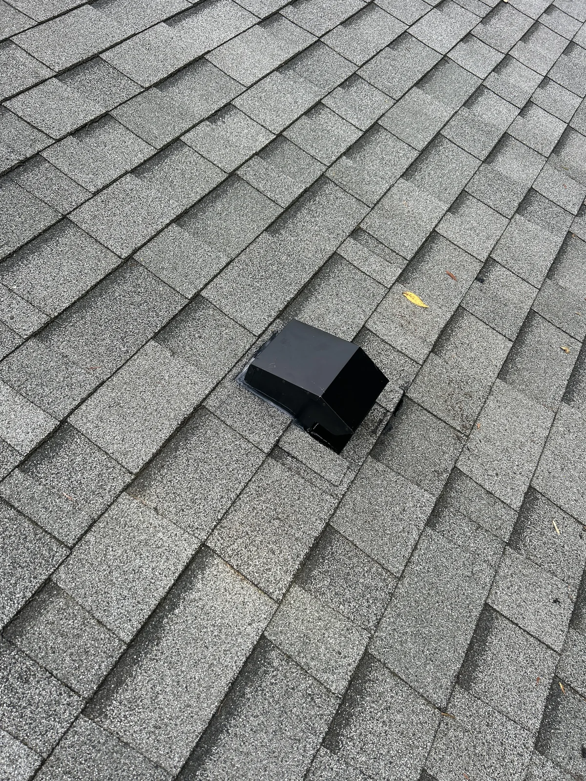 A black rectangular roof vent installed on gray asphalt shingles with a few scattered leaves