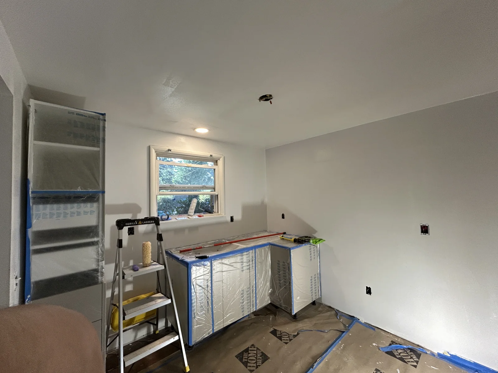 White walls, ceiling light, window, plastic-covered cabinets, ladder, and protective floor covering in a room.