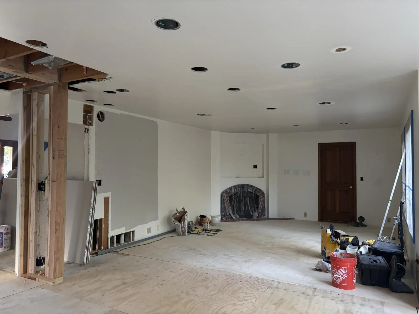 A spacious room with exposed wooden framing, unfinished drywall, plywood flooring, recessed ceiling lights, and construction tools scattered around