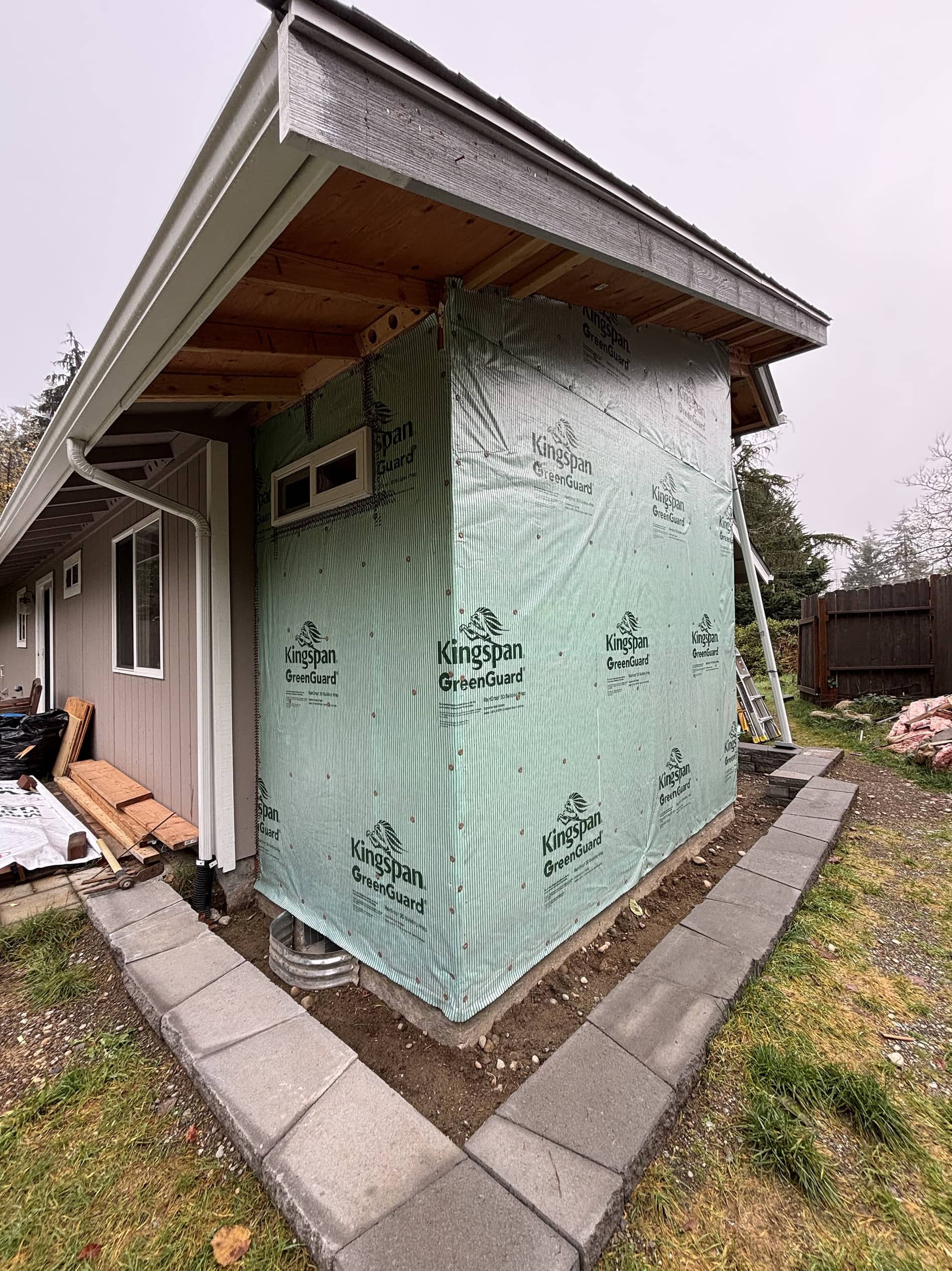 Bathroom addition during construction beside the existing house, with green exterior panels and a small window.