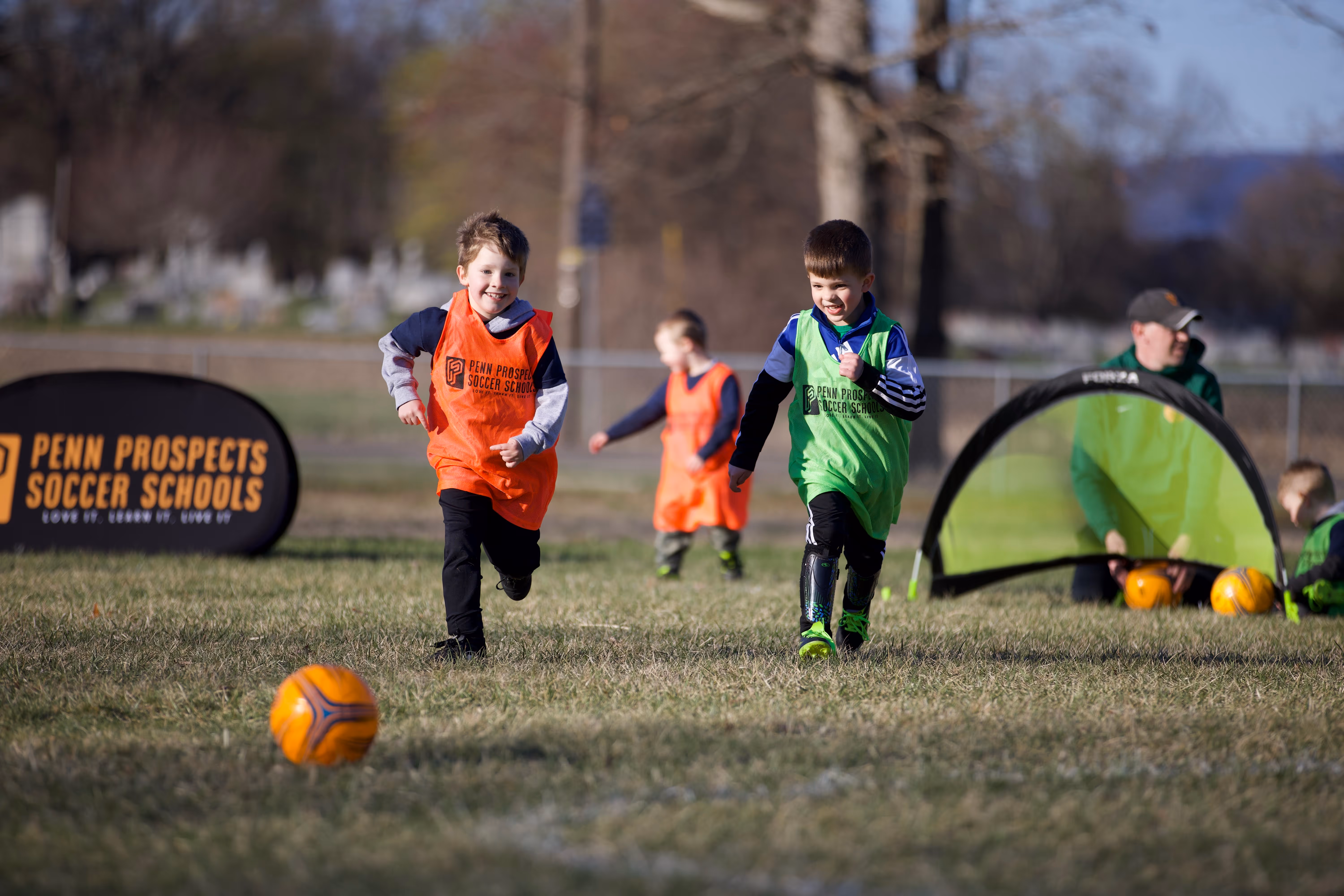 Two young boys joyfully running towards an orange soccer ball on a grassy field during a Penn Prospects Soccer Schools session.