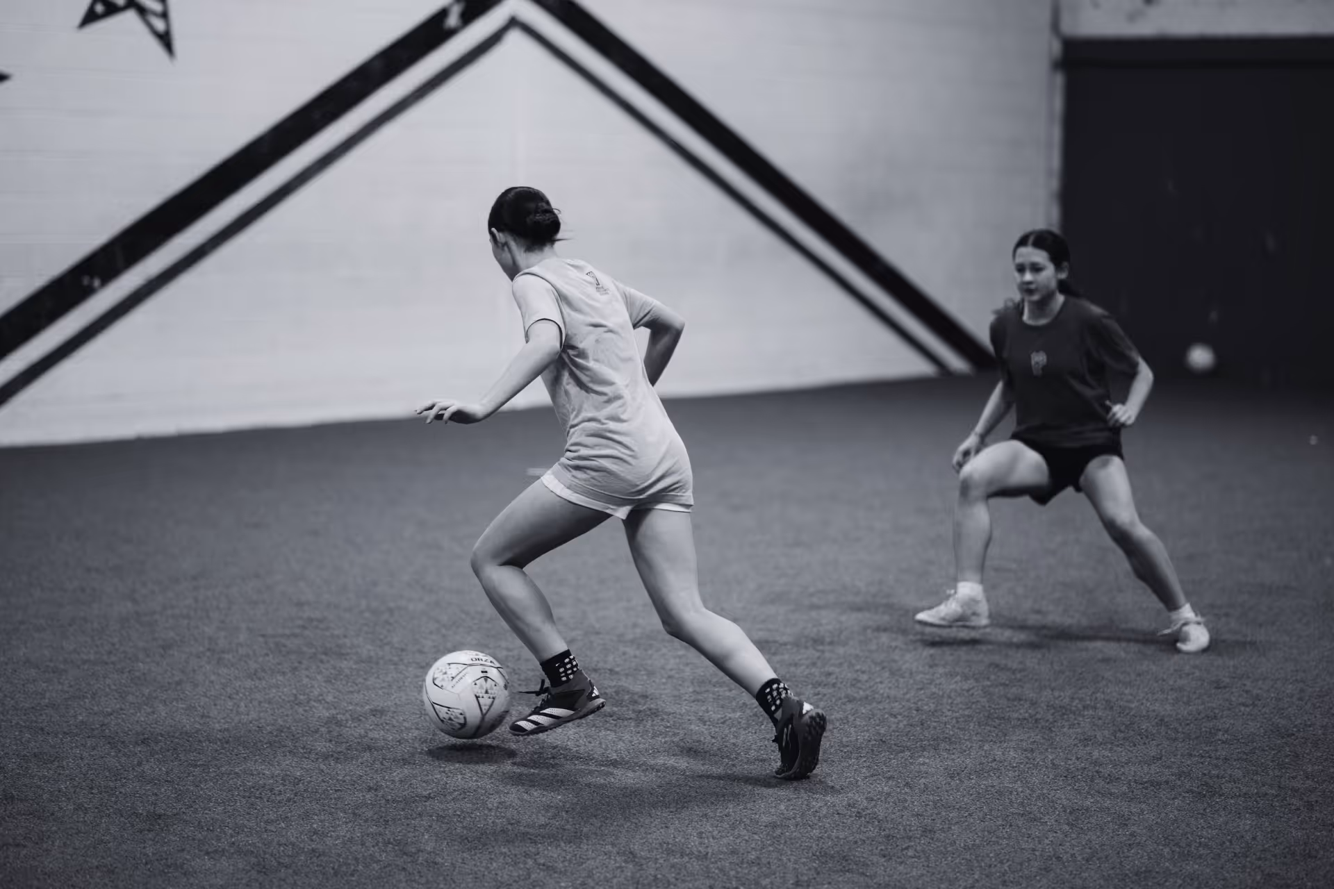 Three girls playing soccer on a grassy field near a goalpost, two in green pinnies and one in an orange pinnie controlling the ball.