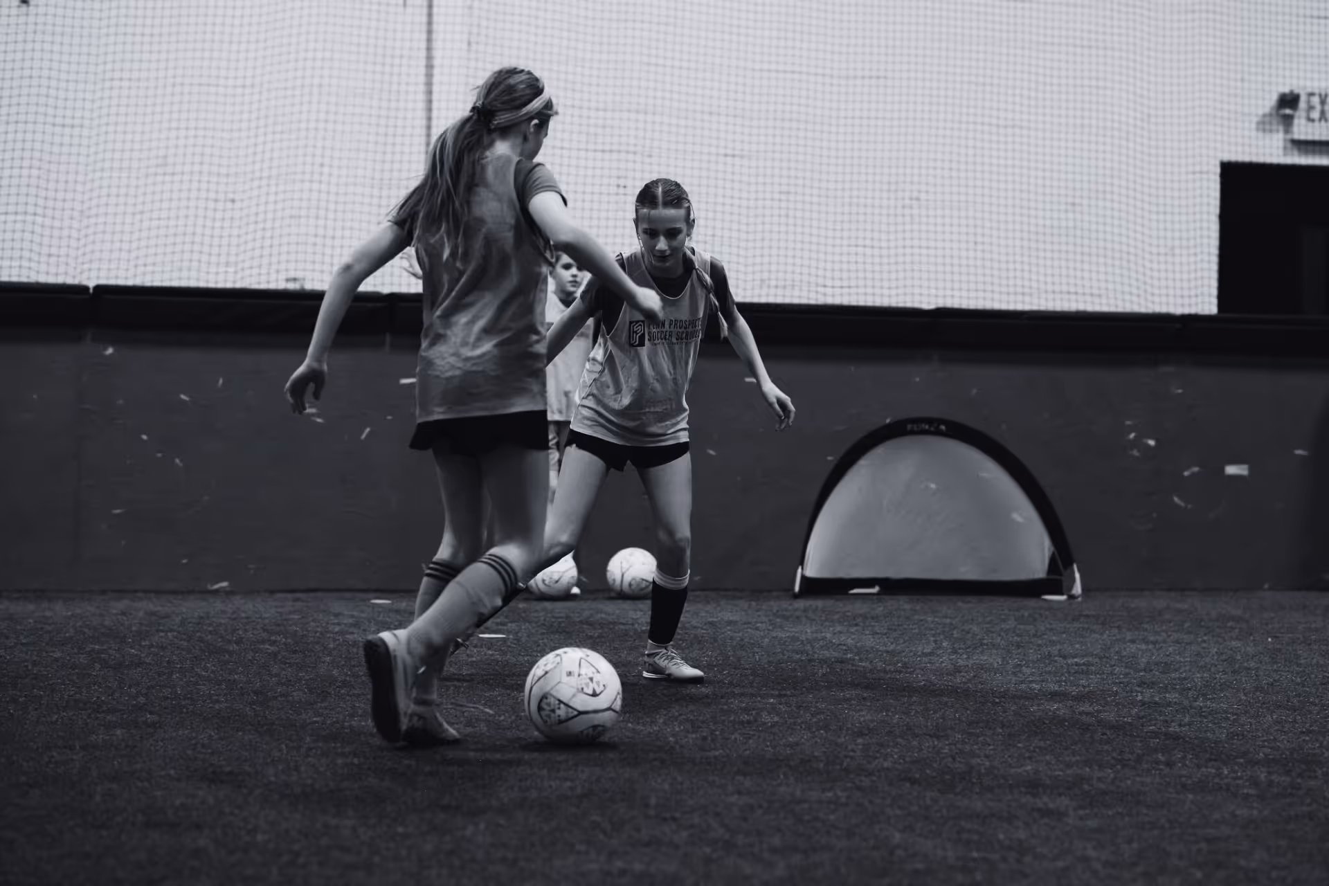 Two young boys playing soccer on a grass field wearing colored bibs, one in orange and the other in green, with a coach nearby.