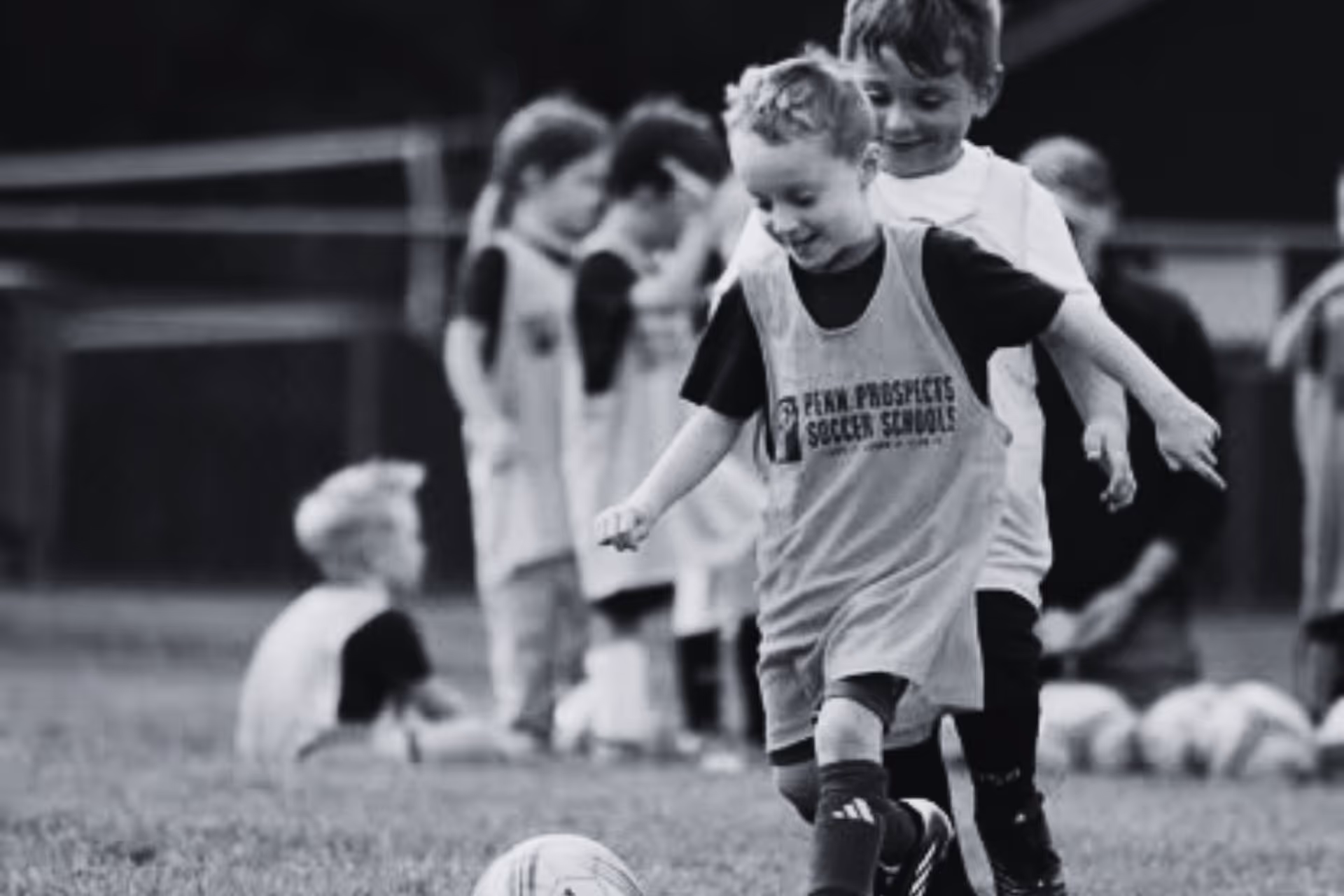 Two young boys playing soccer on a field with other children in the background.