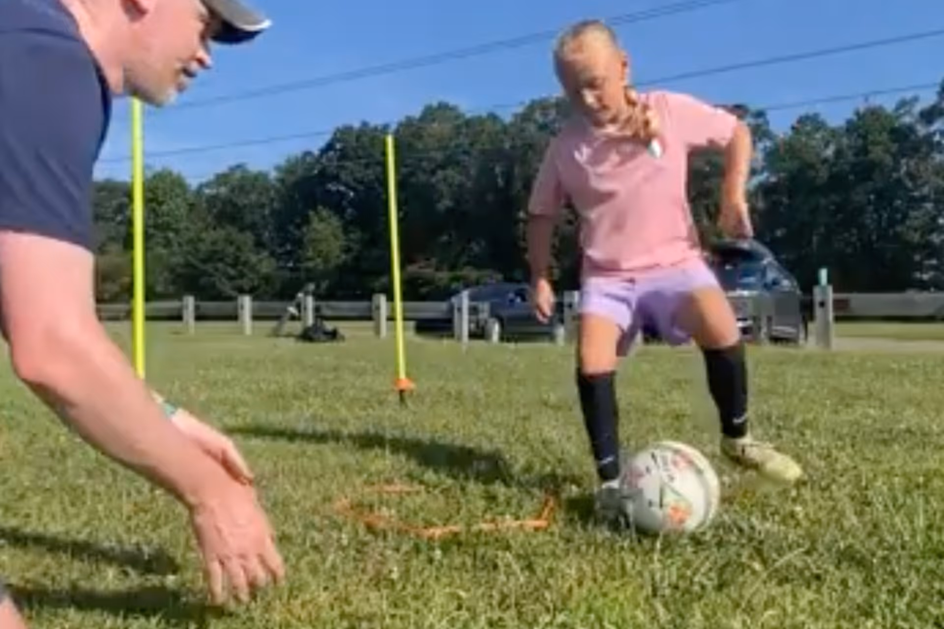Two young boys playing soccer on a grass field wearing colored bibs, one in orange and the other in green, with a coach nearby.