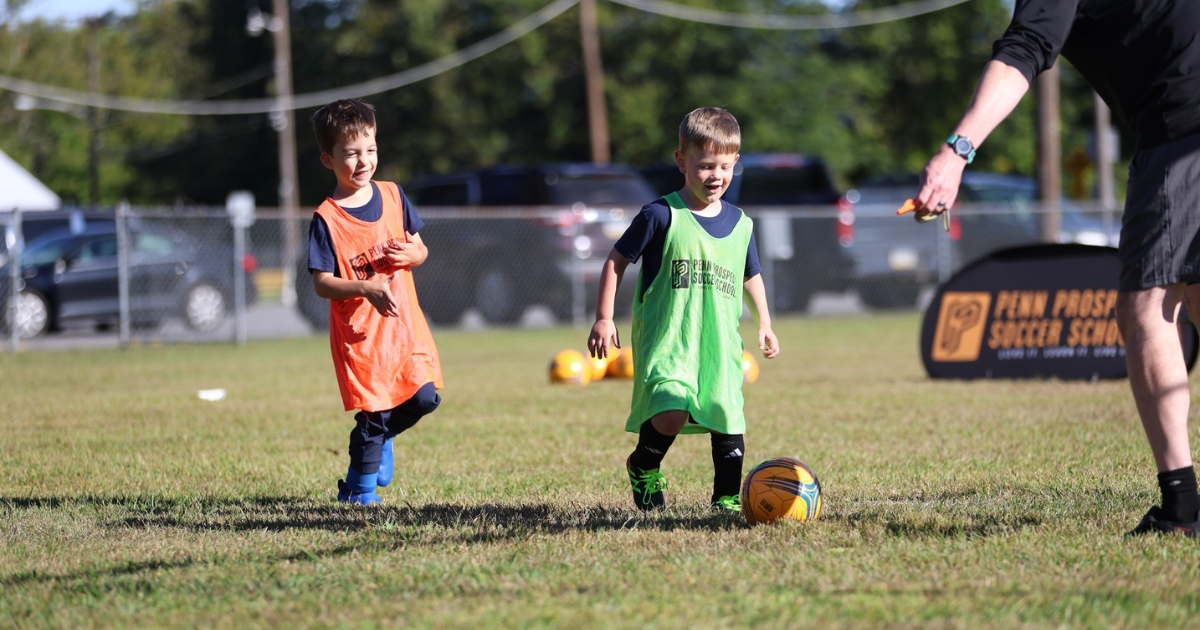 Two young boys playing soccer on a grass field wearing colored bibs, one in orange and the other in green, with a coach nearby.