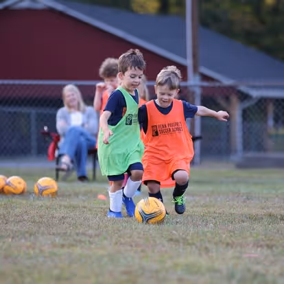 Two young children in green pinnies playing soccer with a yellow ball on grass, supervised by an adult in black.