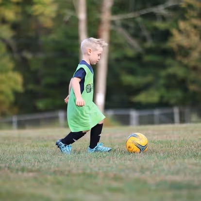 Two young boys playing soccer on a field with other children in the background.