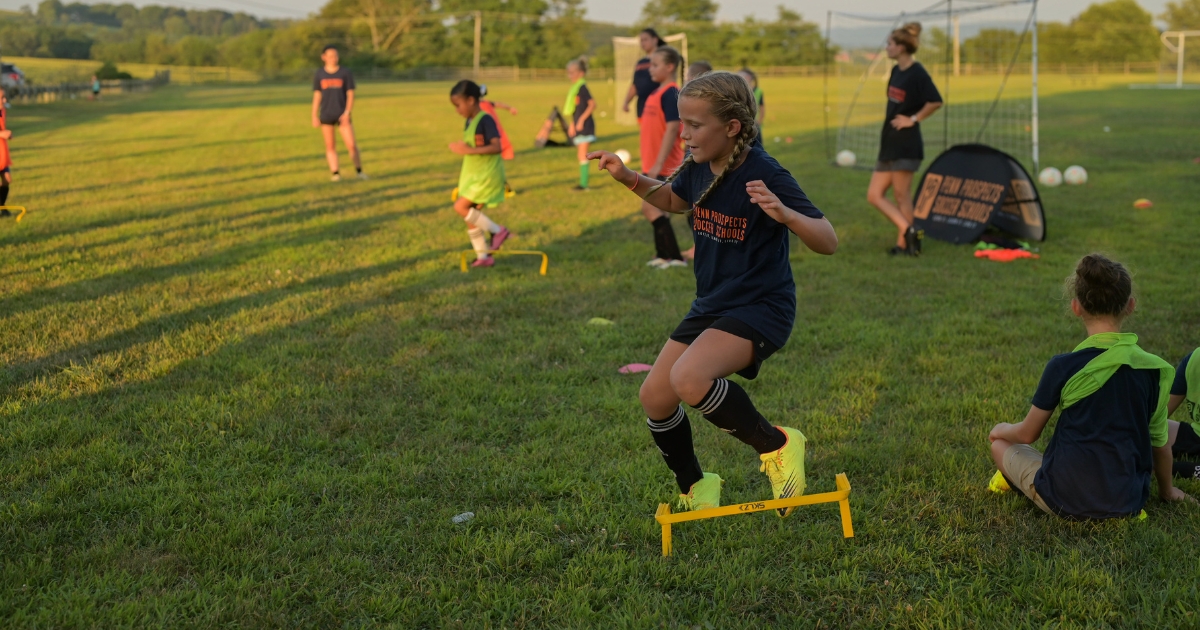 Black and white photo of young children in soccer training vests with soccer balls, raising their hands while their coach kneels beside them on grass.