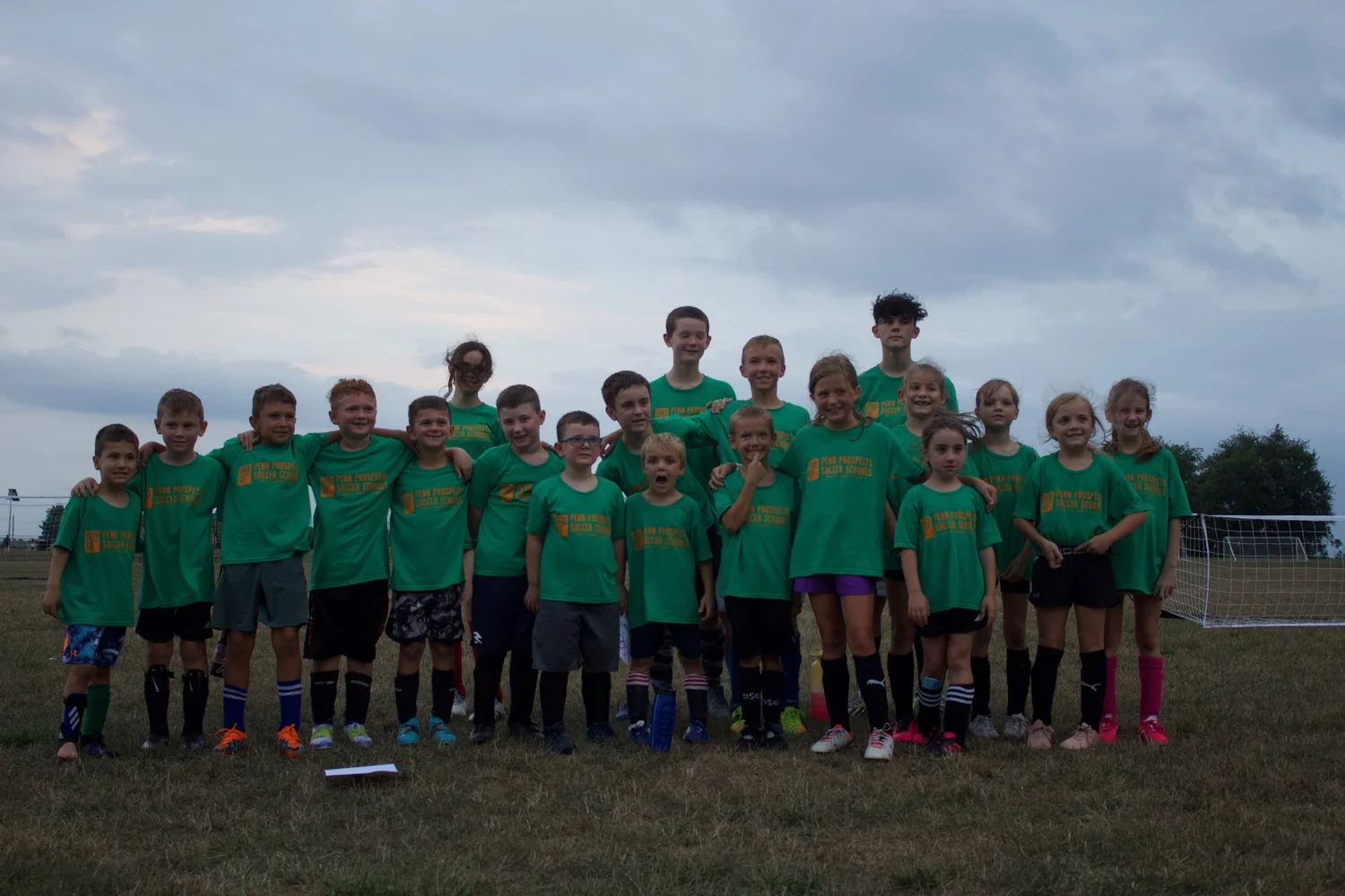 Two young boys in orange and green soccer pinnies playing soccer on a grassy field with an adult partially visible.