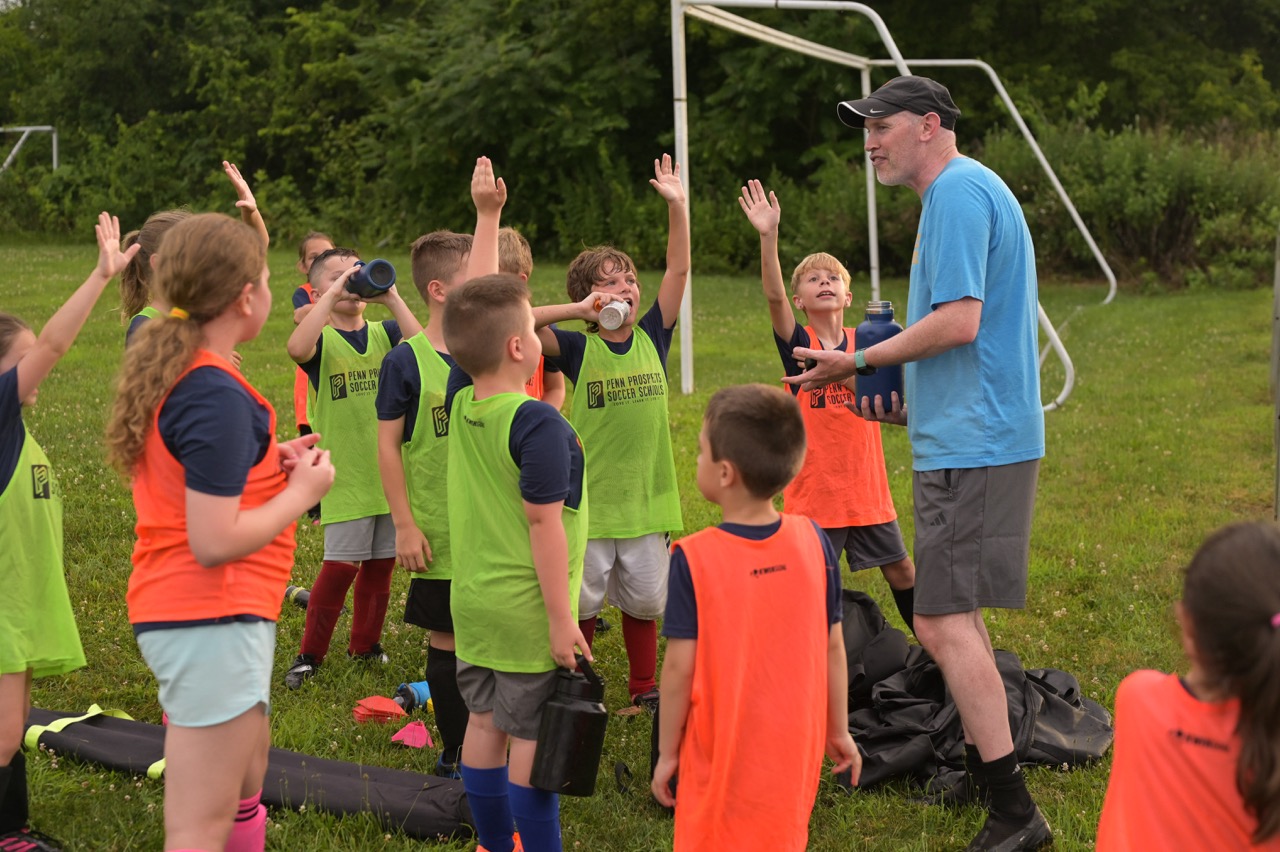 Two young boys playing soccer on a grass field wearing colored bibs, one in orange and the other in green, with a coach nearby.