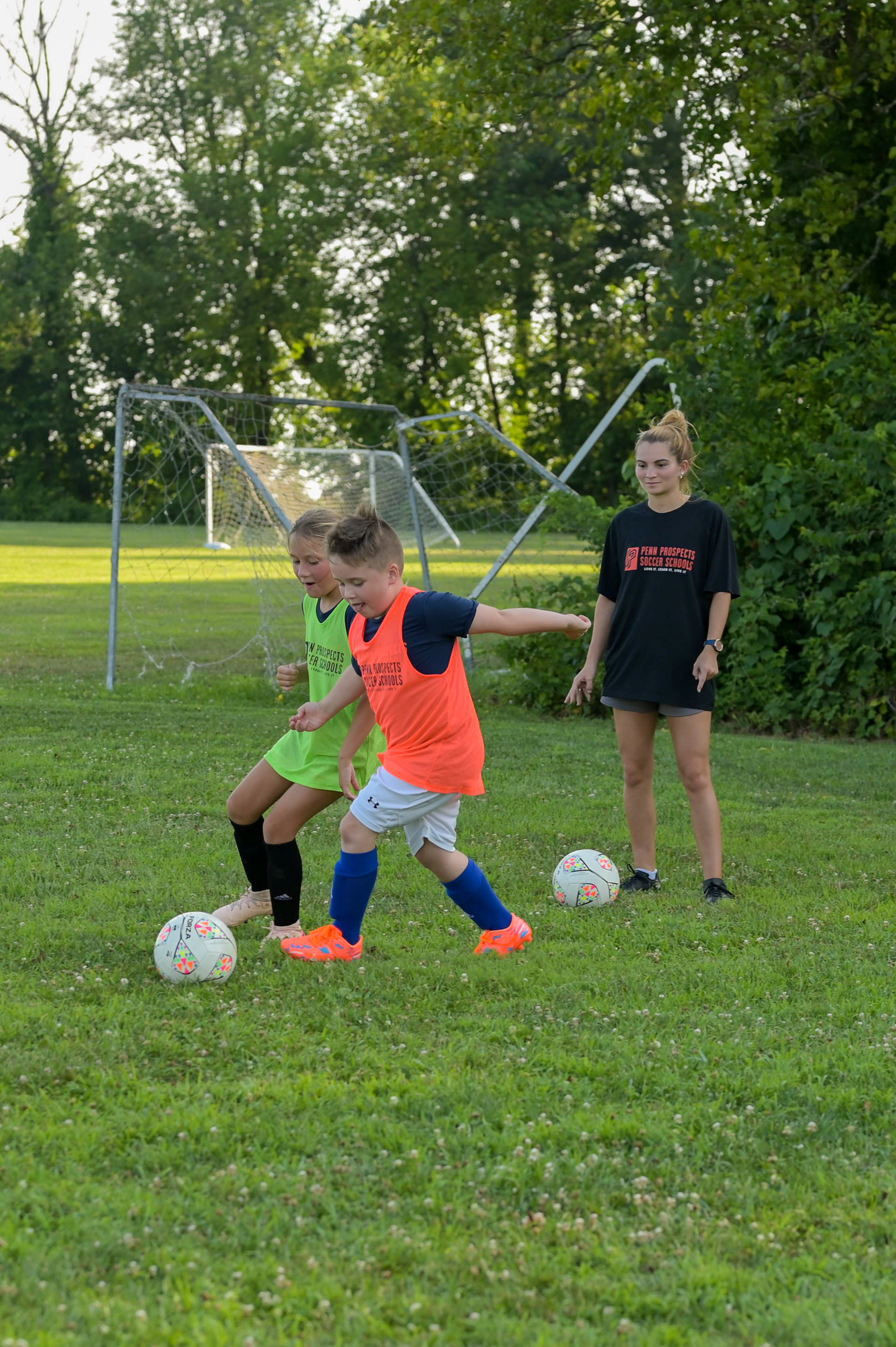 Two young boys playing soccer on a grass field wearing colored bibs, one in orange and the other in green, with a coach nearby.