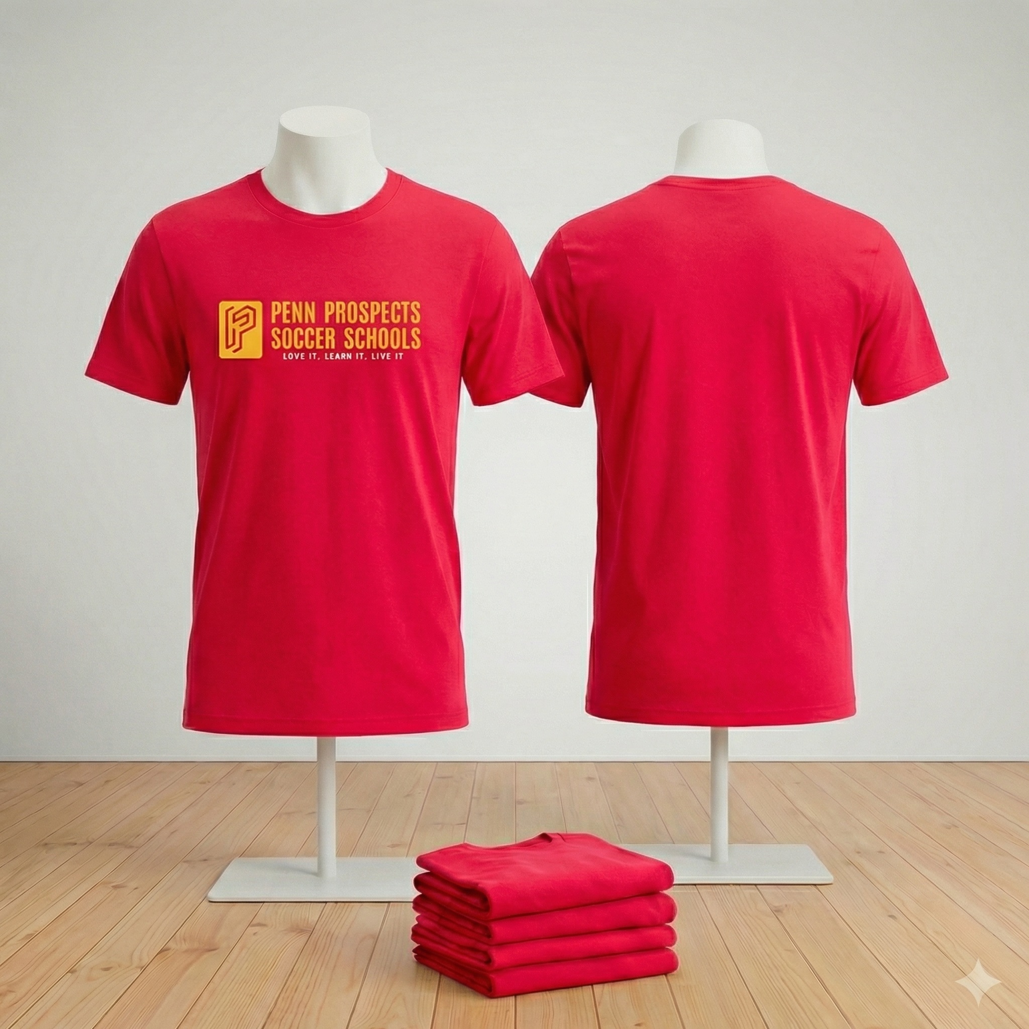 Red T-shirts displayed on mannequins, front showing Penn Prospects Soccer Schools logo and slogan, back plain, with a stack of folded red T-shirts on a wooden floor.