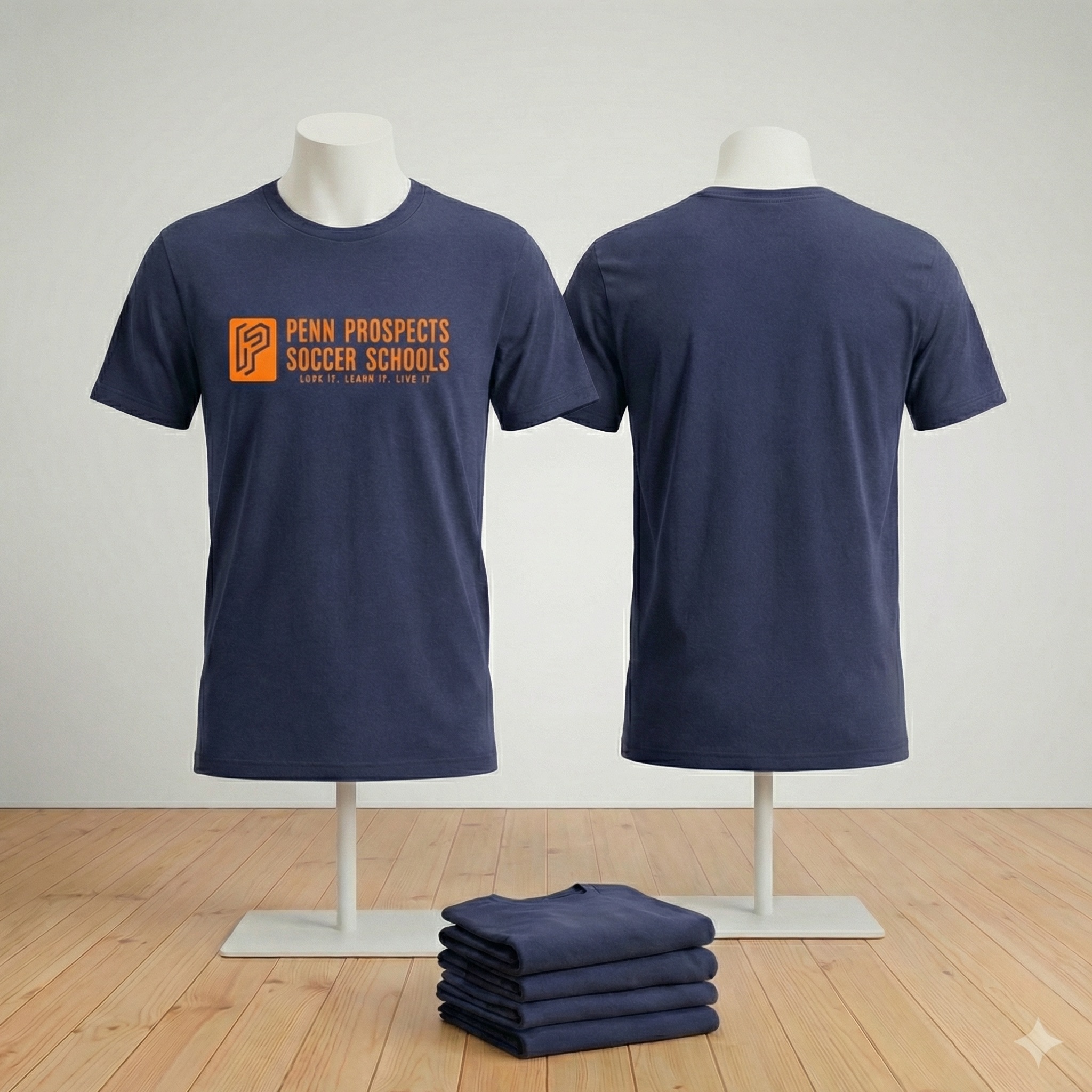 Navy blue t-shirt displayed front and back on mannequins with orange text reading 'PENN PROSPECTS SOCCER SCHOOLS' and slogan below, with folded shirts stacked on the floor.
