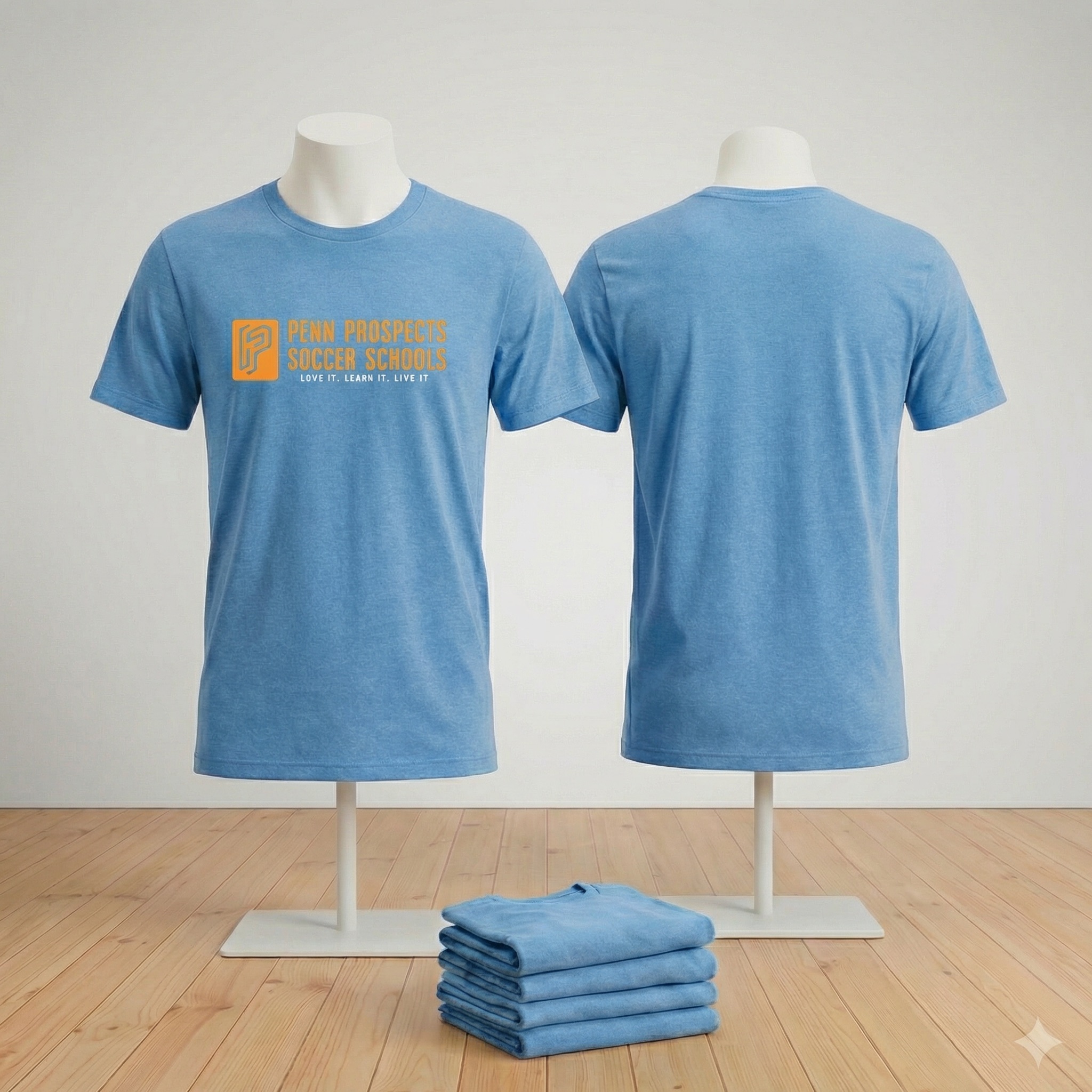 Front and back view of a blue t-shirt on mannequins with 'Penn Prospects Soccer Schools' logo and slogan in orange on the front, with a stack of folded blue t-shirts in front.