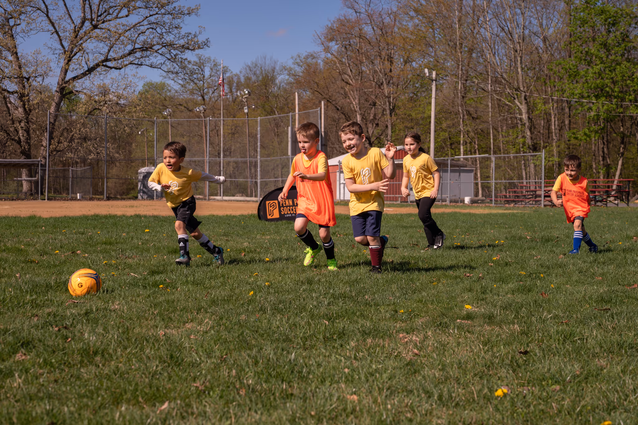 Two young children in green pinnies playing soccer with a yellow ball on grass, supervised by an adult in black.