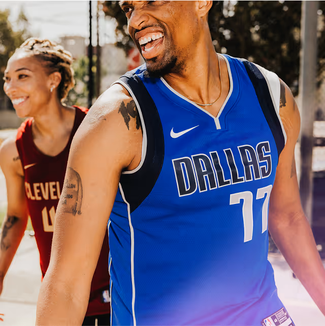 A man wearing a blue Dallas Mavericks #77 basketball jersey smiles as he walks alongside a friend.