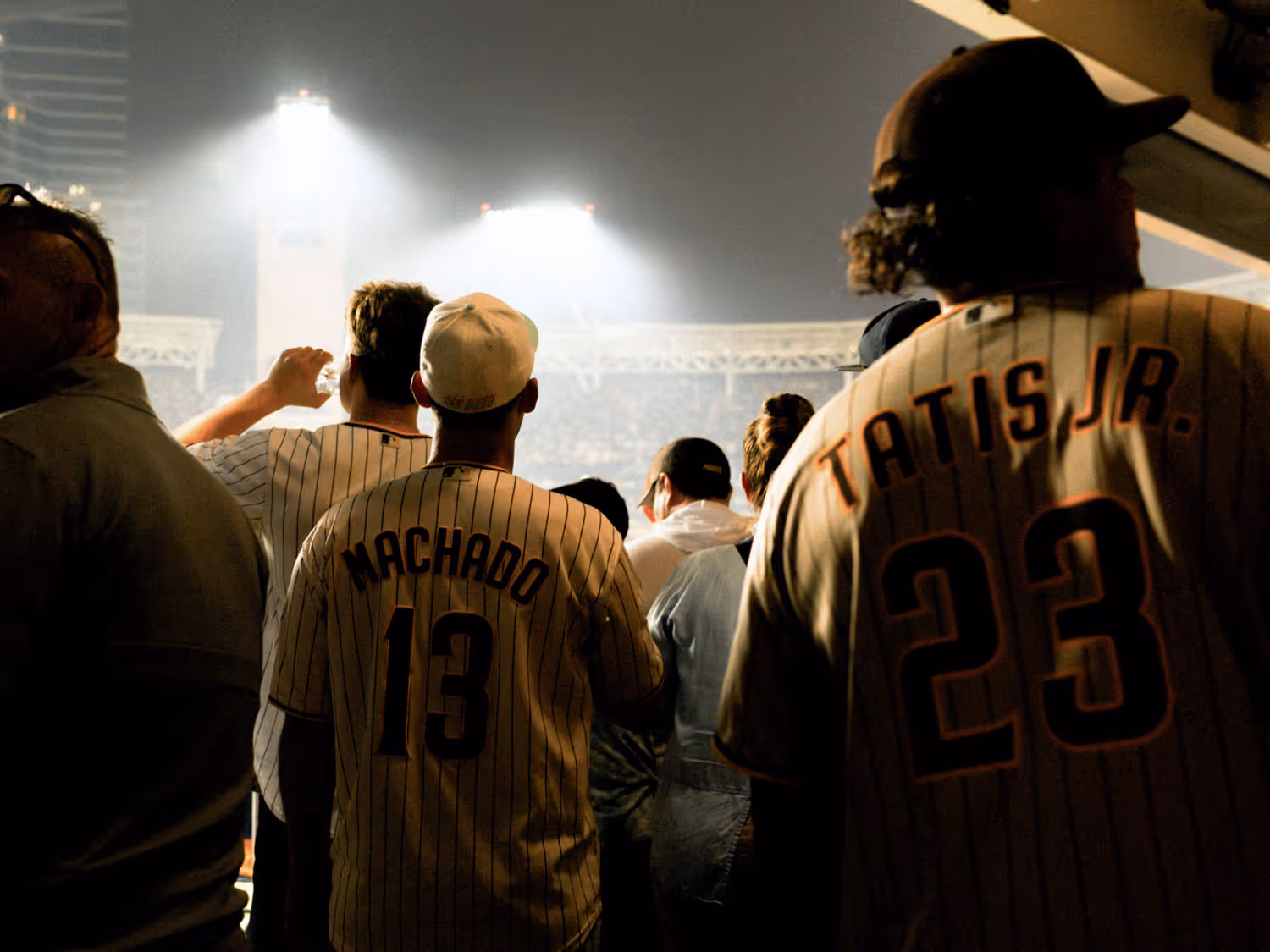 A spectator wearing a #8 Pence baseball jersey walking down a stadium concourse at night, with other fans and bright stadium lights in the background.