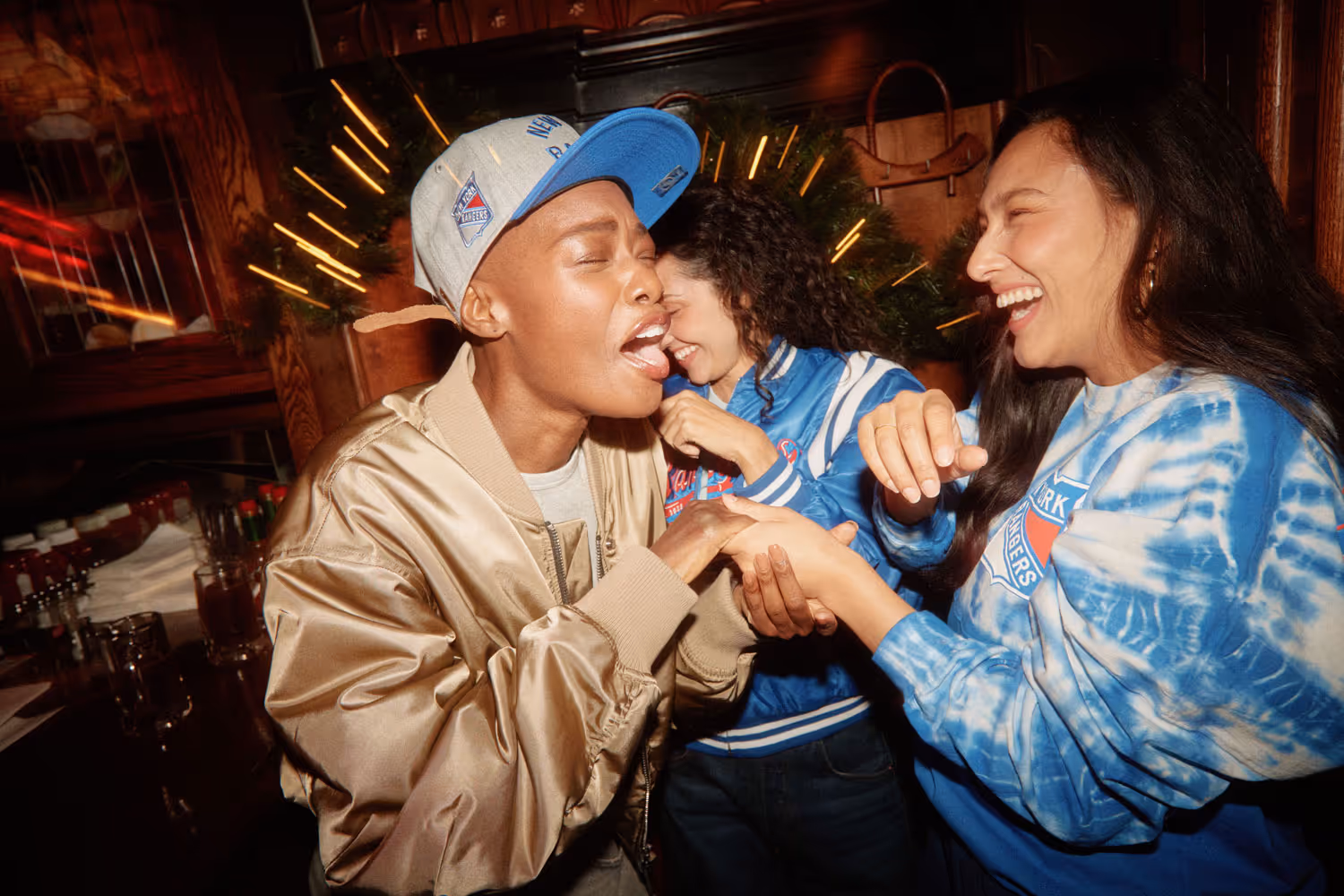 Three women laughing together in a dimly-lit bar; one playfully pretends to bite another’s hand while a third smiles behind them under neon lights.