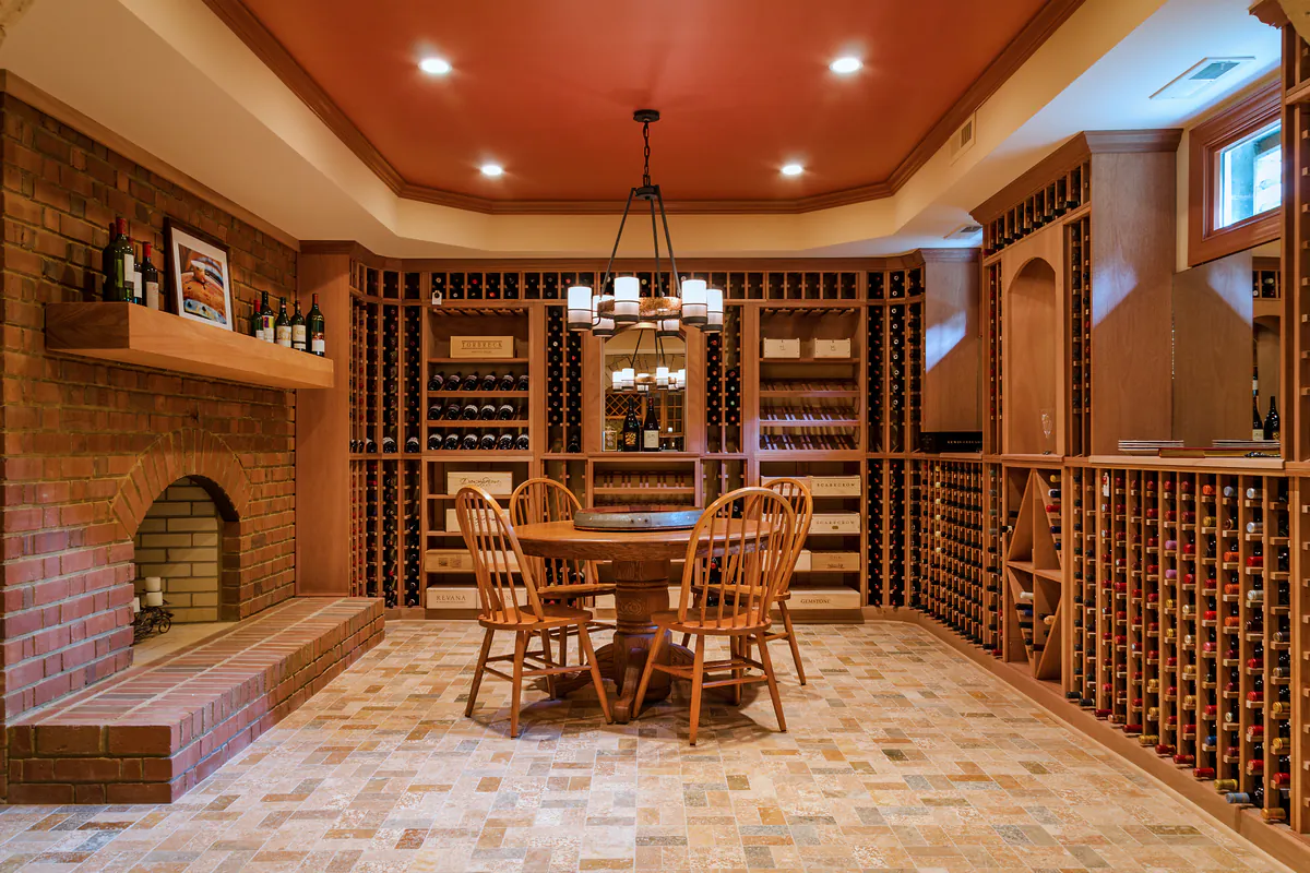 Wine cellar with warm wood finishes, custom built-in wine racks, and a central round table with four chairs. Features a brick fireplace, soft overhead lighting, and a terracotta ceiling, creating an inviting space for wine tasting and entertaining.