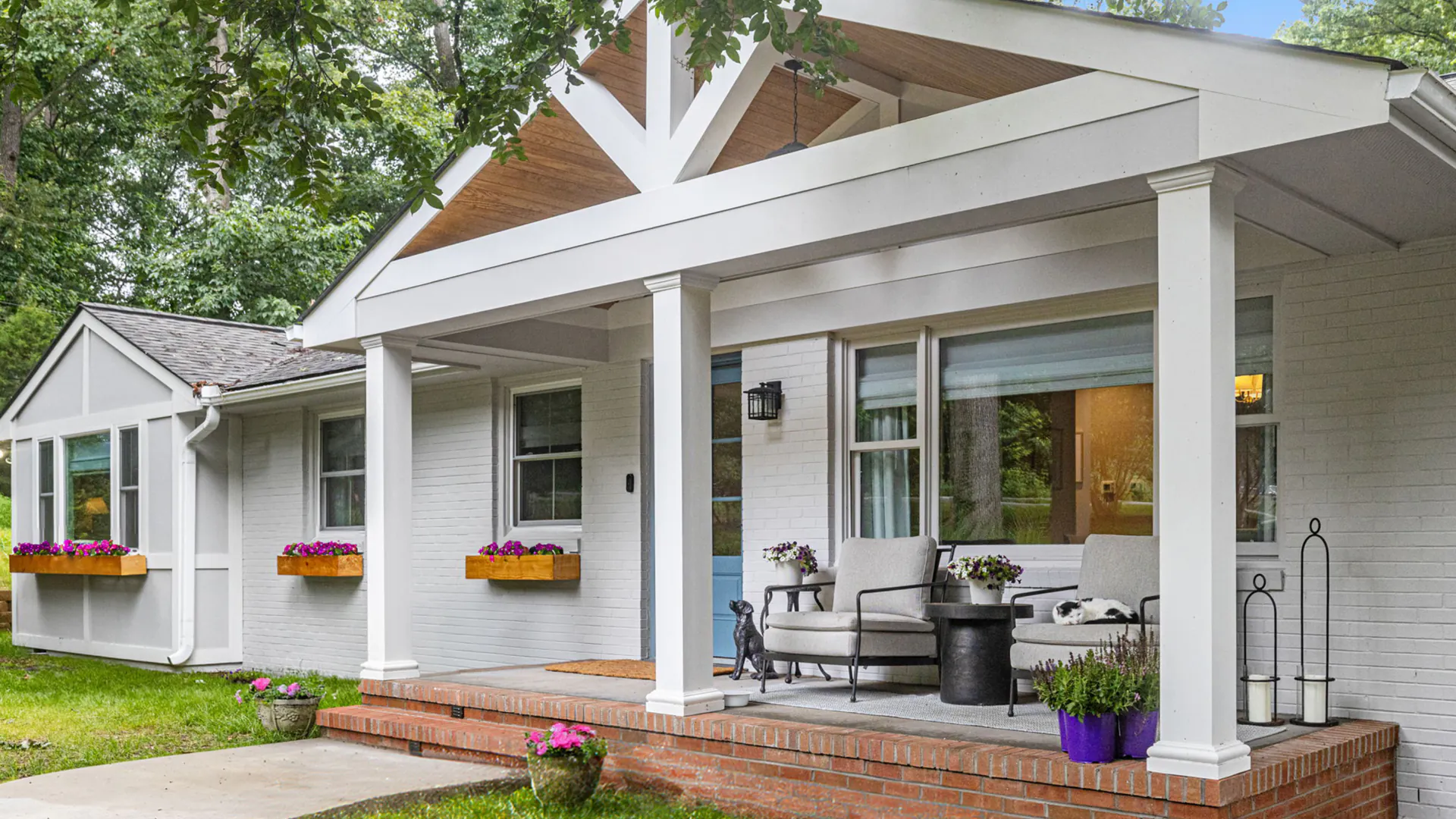 Covered front porch addition with white columns and brick foundation, enhancing the curb appeal of a painted brick ranch home. Features include window flower boxes, cozy outdoor seating, and decorative planters for a welcoming entry.