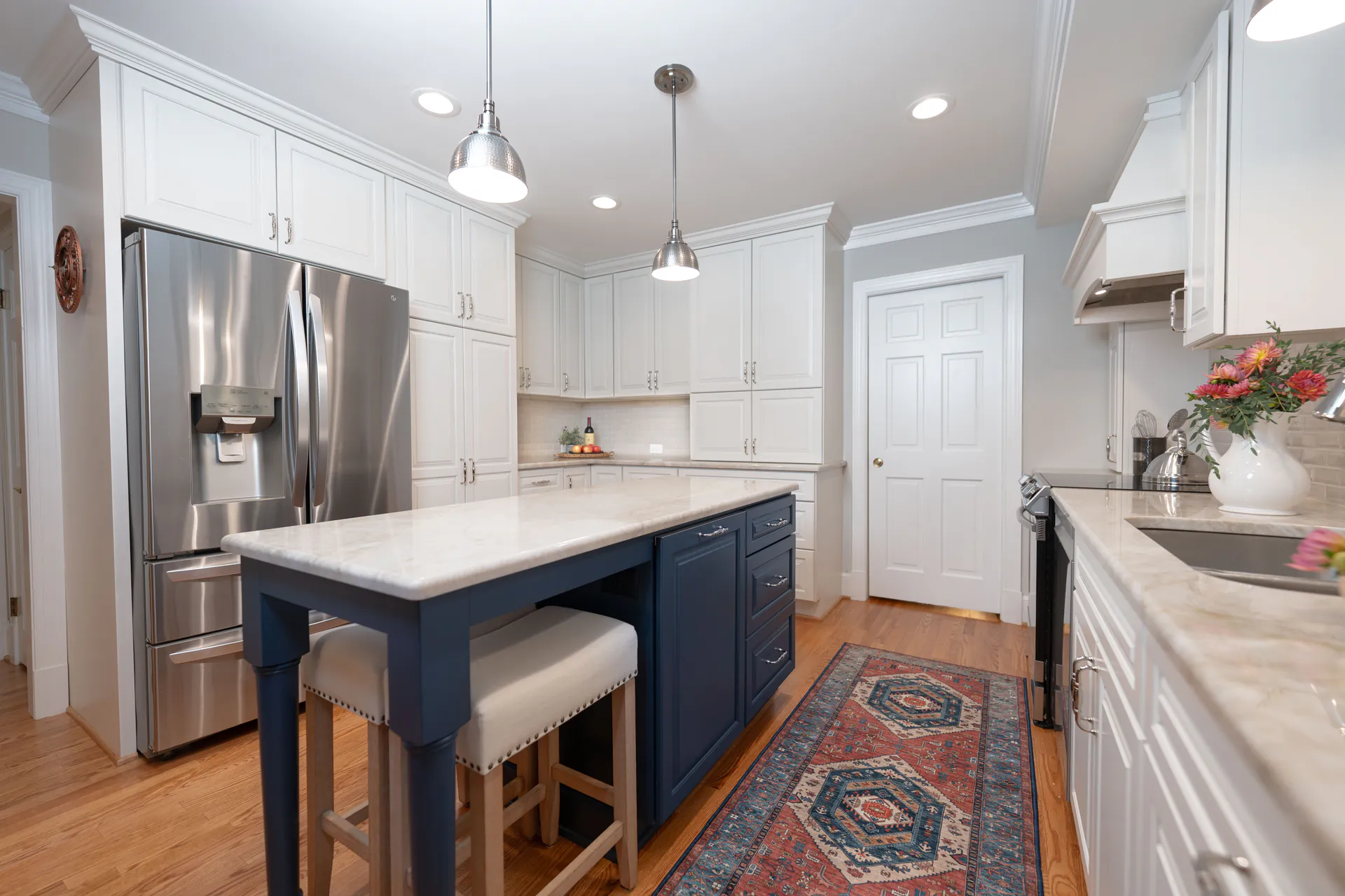 Bright kitchen with white cabinetry, a navy island with seating, stainless steel refrigerator, pendant lighting, and a patterned runner on hardwood floors.