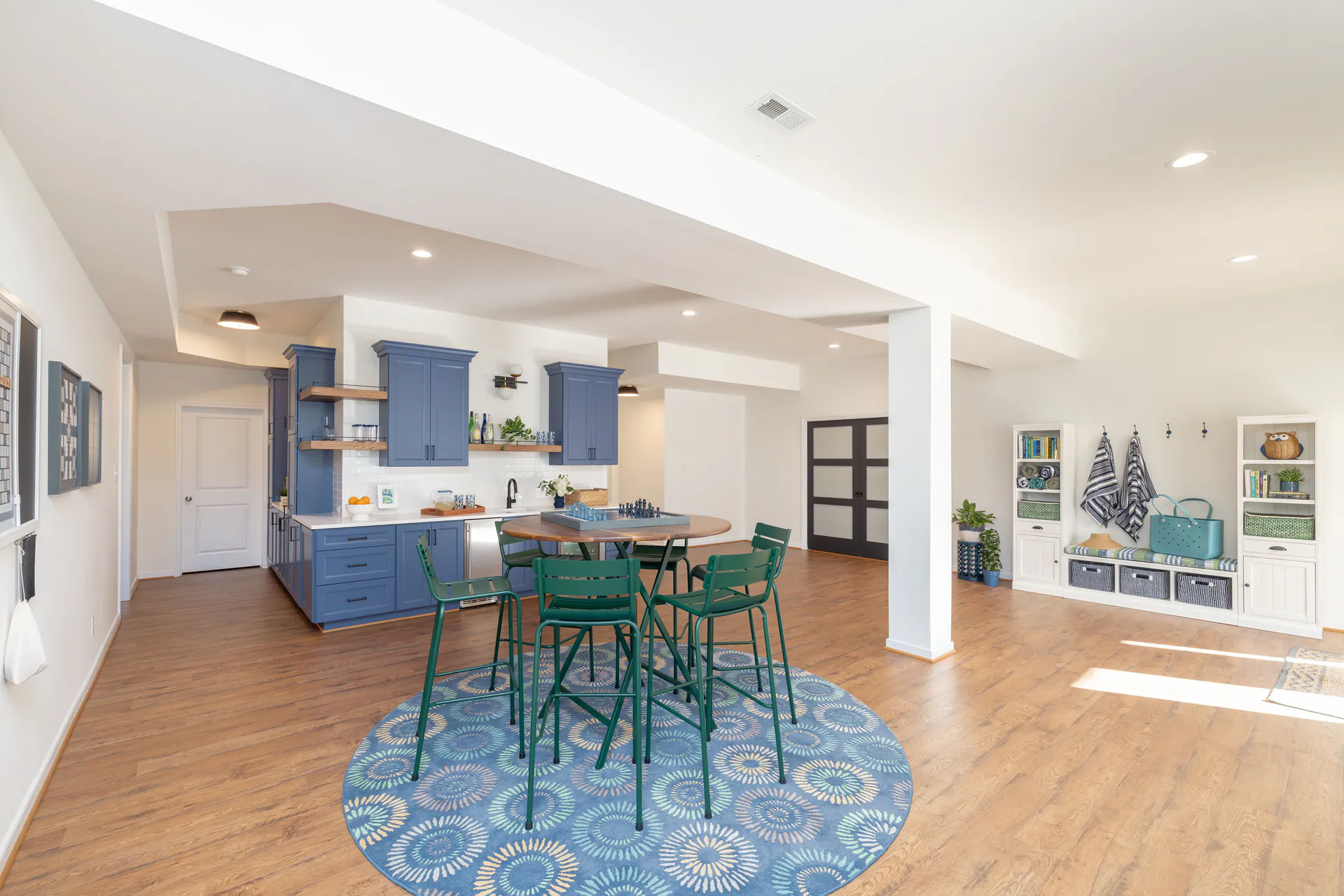 Open-concept finished basement featuring a blue kitchenette with custom cabinetry, wood-look flooring, a round game table with seating, recessed lighting, and built-in storage creating a bright, functional lower-level living space.