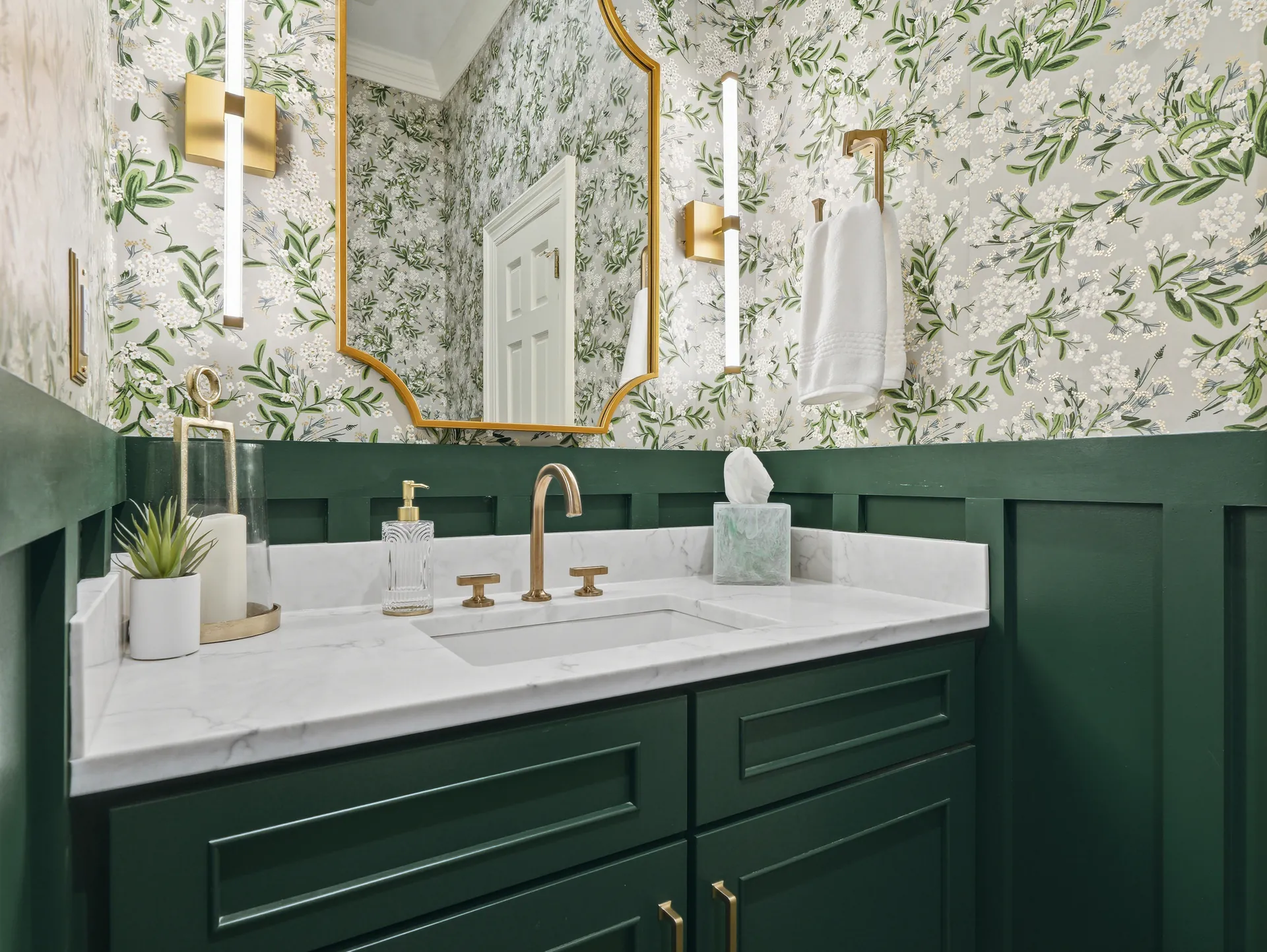 Walk-in shower with glossy emerald green subway tile walls, pebble tile flooring, and a frameless glass panel. Features include a brushed gold rainfall shower head with handheld fixture, built-in niche, grab bar, and a wooden bench. Natural light filters through a window with white plantation shutters, and botanical wallpaper accents the upper wall.
