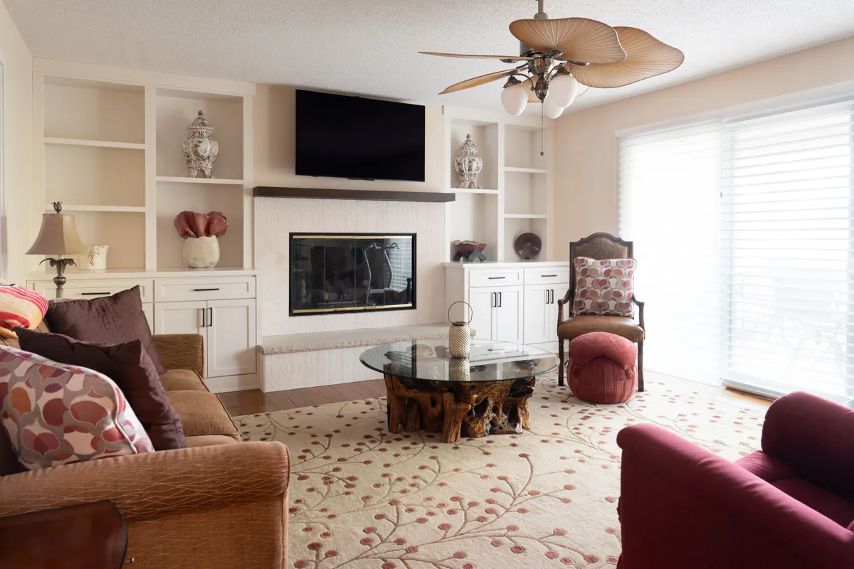 Bright living room remodel featuring custom white built-in shelving and cabinetry surrounding a modern fireplace with a mounted TV above. The space includes warm wood flooring, a large patterned area rug, contemporary seating with red and brown accents, a sculptural glass coffee table, and expansive sliding glass doors that fill the room with natural light.