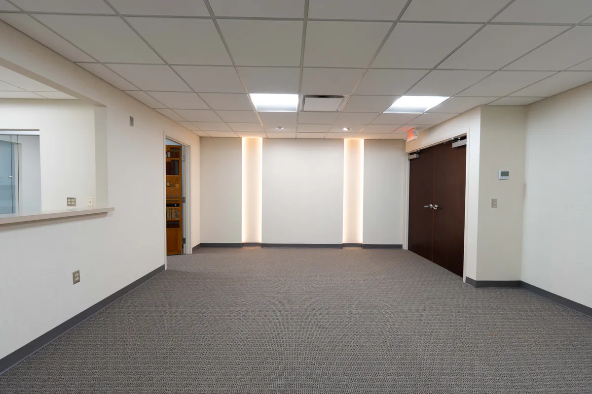 Newly remodeled commercial office interior with neutral walls, patterned carpet flooring, recessed accent lighting, and dark wood double doors. The open lobby or waiting area features a drop ceiling with bright overhead lighting and a clean, modern design.