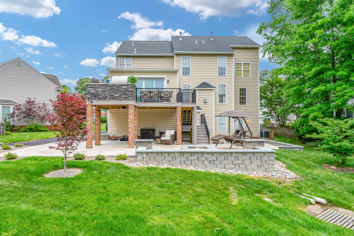 Rear exterior view of a multi-level outdoor living renovation featuring a covered patio with a fireplace lounge below, an elevated deck with modern railing and stone-clad privacy wall above, and a spacious backyard with landscaped retaining walls, lounge seating, and mature trees surrounding the property.