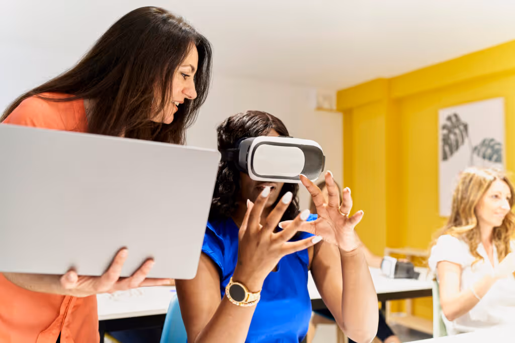 teacher in classroom holding laptop standing next to female student with hand on her man while instructing her on how to use virtual reality goggles