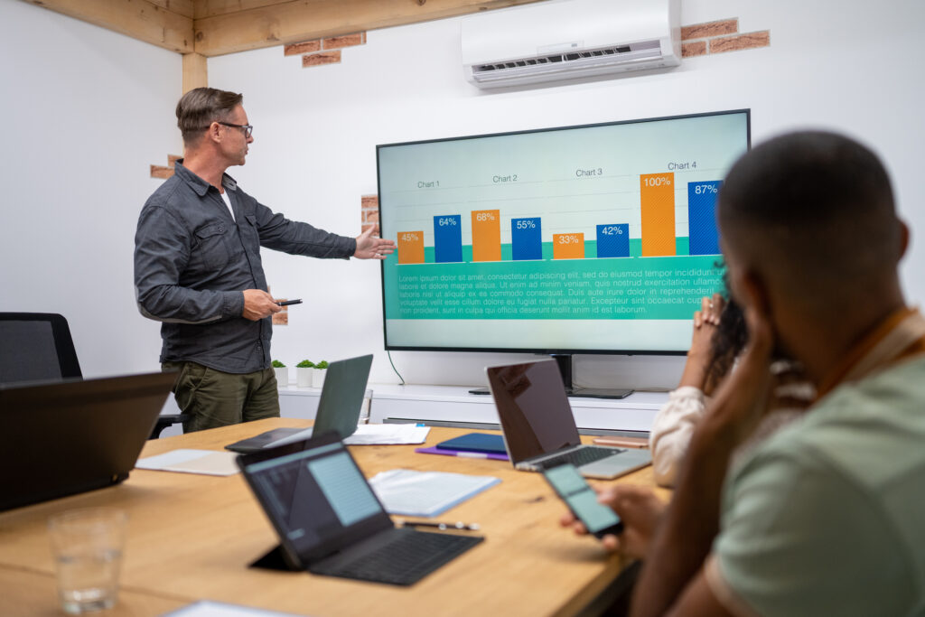 Businessman pointing at TV screen while discussing business plan with team during meeting. An example of what can be created using a presentation generator ai tool. 