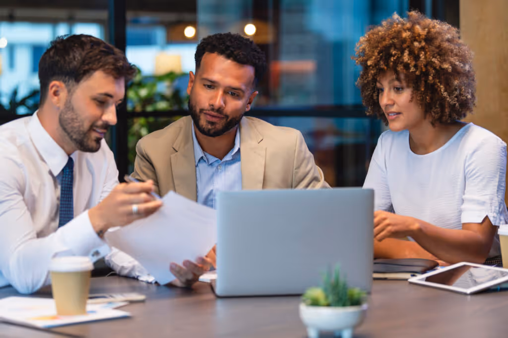 Three sales people meeting and looking at a laptop and a document. There is paperwork and other technology on the table, formal business wear.