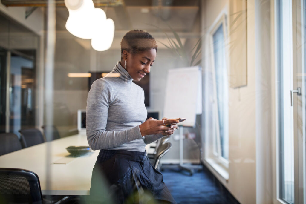Mid adult businesswoman using cell phone. Confident female professional is working in new office standing in conference room reading text message on her mobile phone.