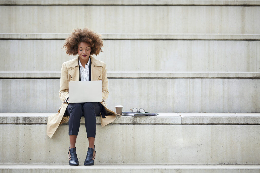 Young businesswoman using laptop while sitting on steps. Full length of female professional working on staircase. She may be using a presentation generator ai to speed up her presentation creation process. 
