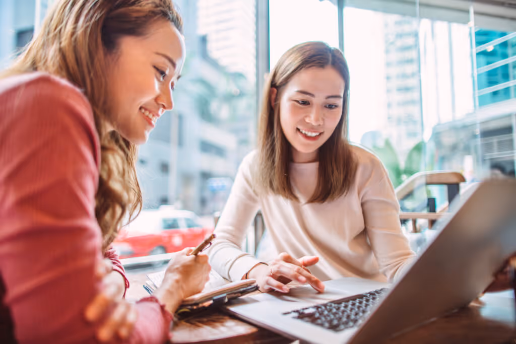 Two young cheerful businesswomen working on laptop computer together on laptop computer in cafe. One of them is jotting down notes during the meeting.