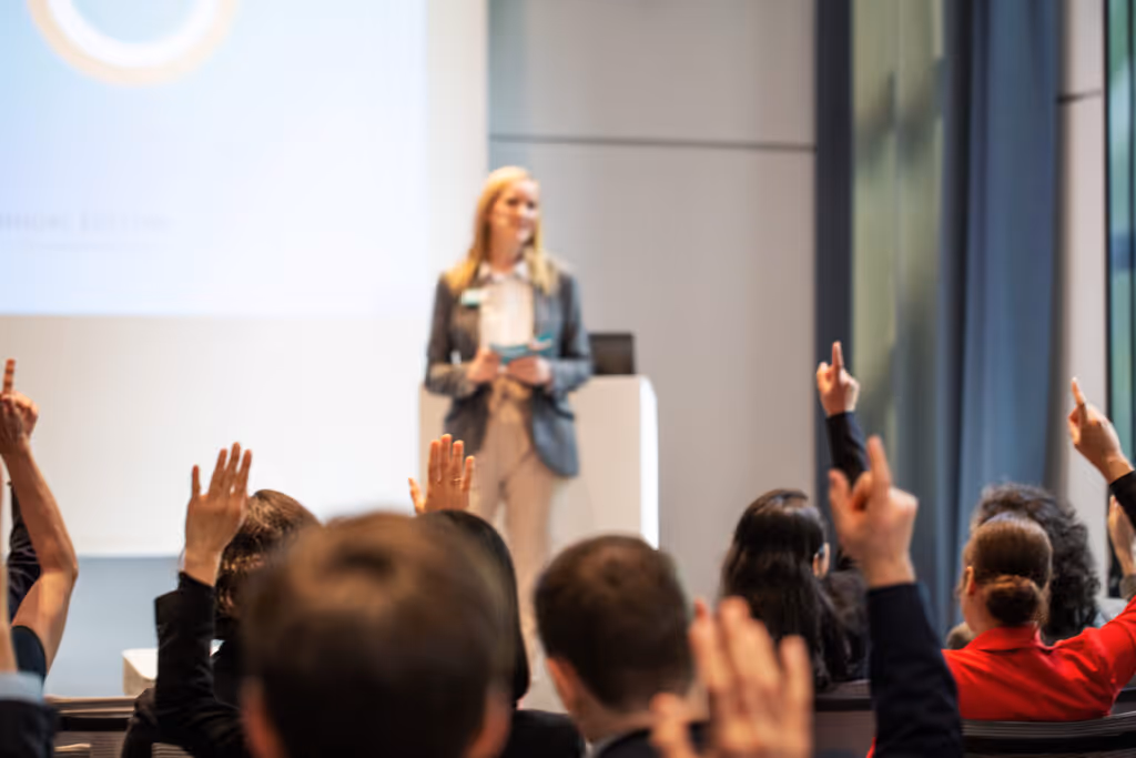 Business people raising hands to ask questions during a presentation. Woman giving a good presentation with people sitting in front raising hands at convention center.