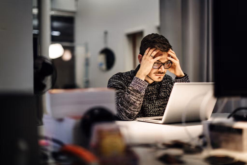 Young casual businessman wearing glasses is sitting in front of his notebook holding his head pondering over his work. Office equipment and another computer is in front of him.