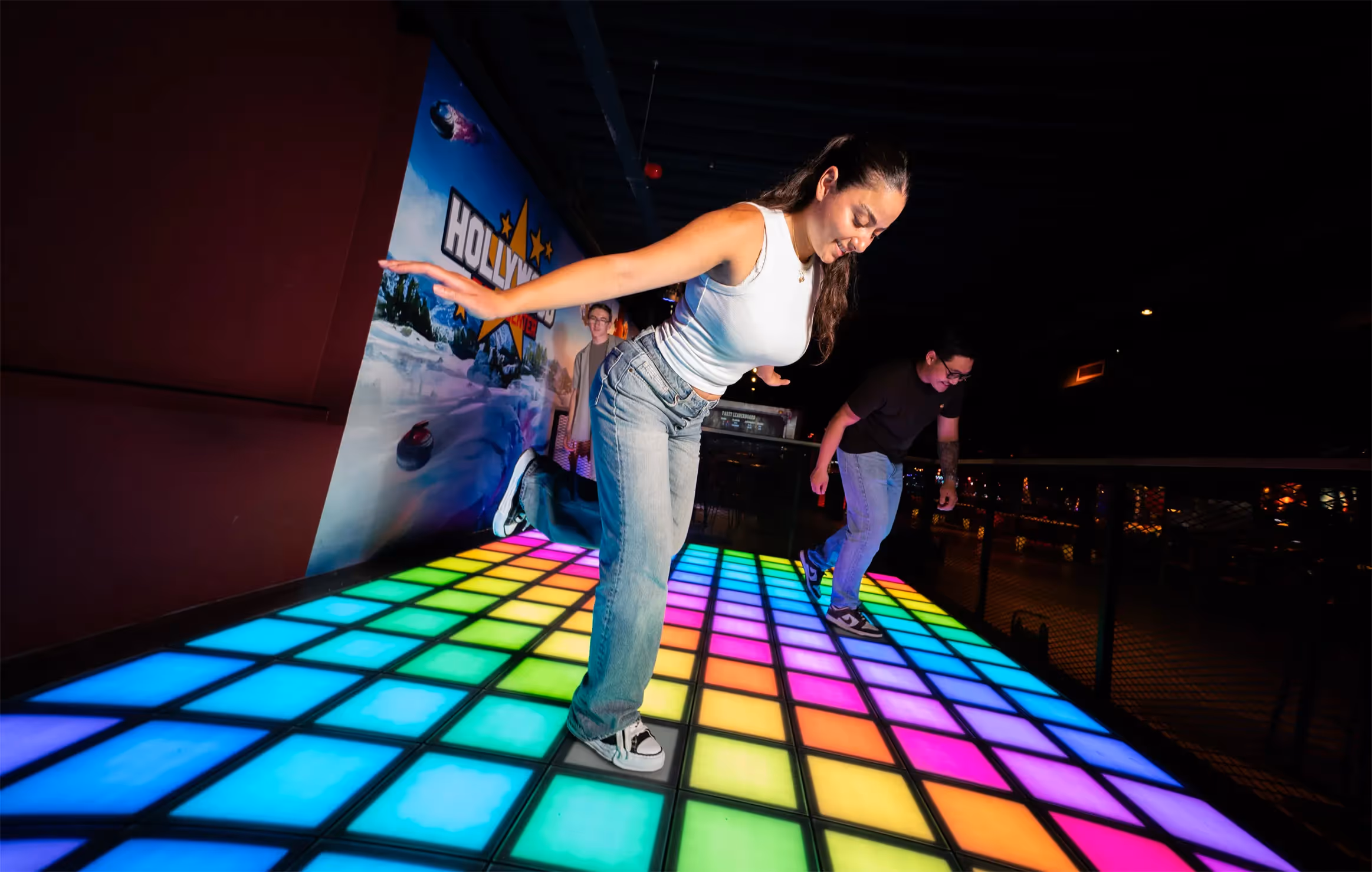 Two people balancing and playing a game on a colorful lighted dance floor in a dark indoor setting.