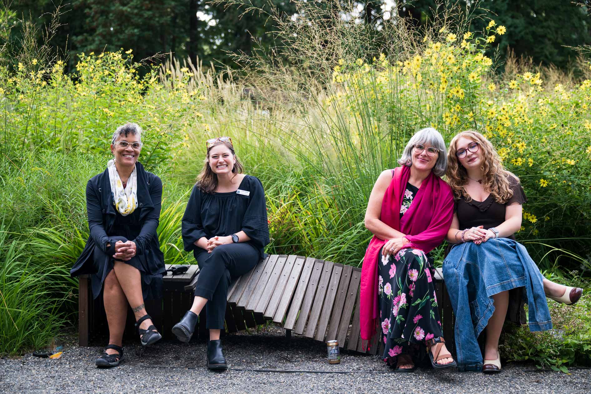 A joyful group of four women sits on a wooden bench, smiling and laughing in a lush garden at the Feast in the Garden event in Portland, Oregon. The women are all looking at the camera, sharing a moment of genuine connection and happiness. The background is filled with tall grasses and yellow wildflowers, capturing the warmth and community spirit of the event.