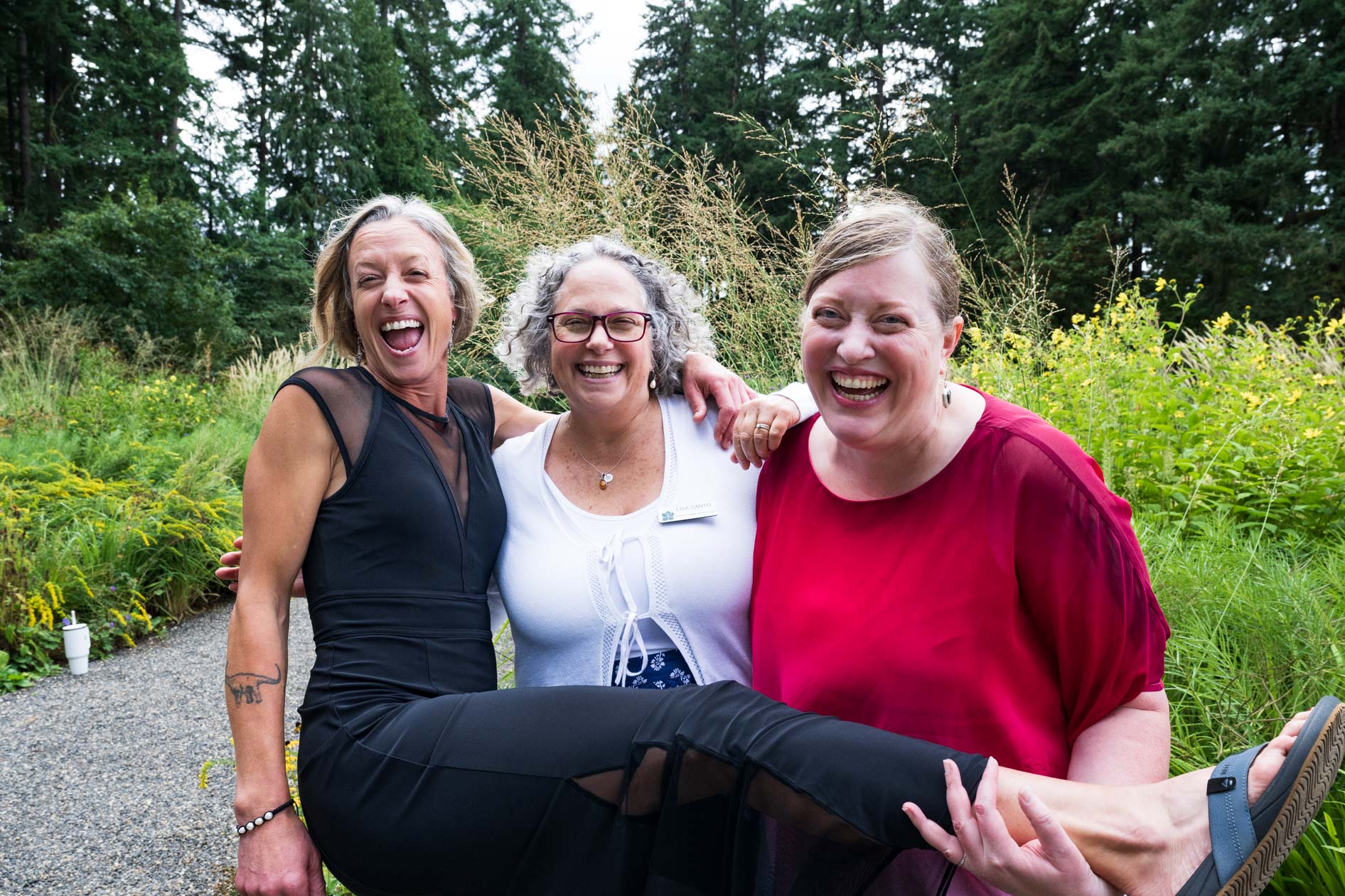 A hilarious and candid moment between three women who are laughing and embracing, with one of them playfully lifting the other's leg. They are standing in a lush garden at the Feast in the Garden event, showcasing the genuine joy of the evening.