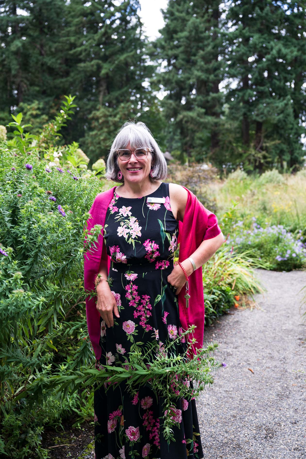 A woman in a floral dress and pink shawl smiles, radiating confidence at the Feast in the Garden event in SE Portland. She stands on a path surrounded by the lush greenery of Leach Botanical Garden.
