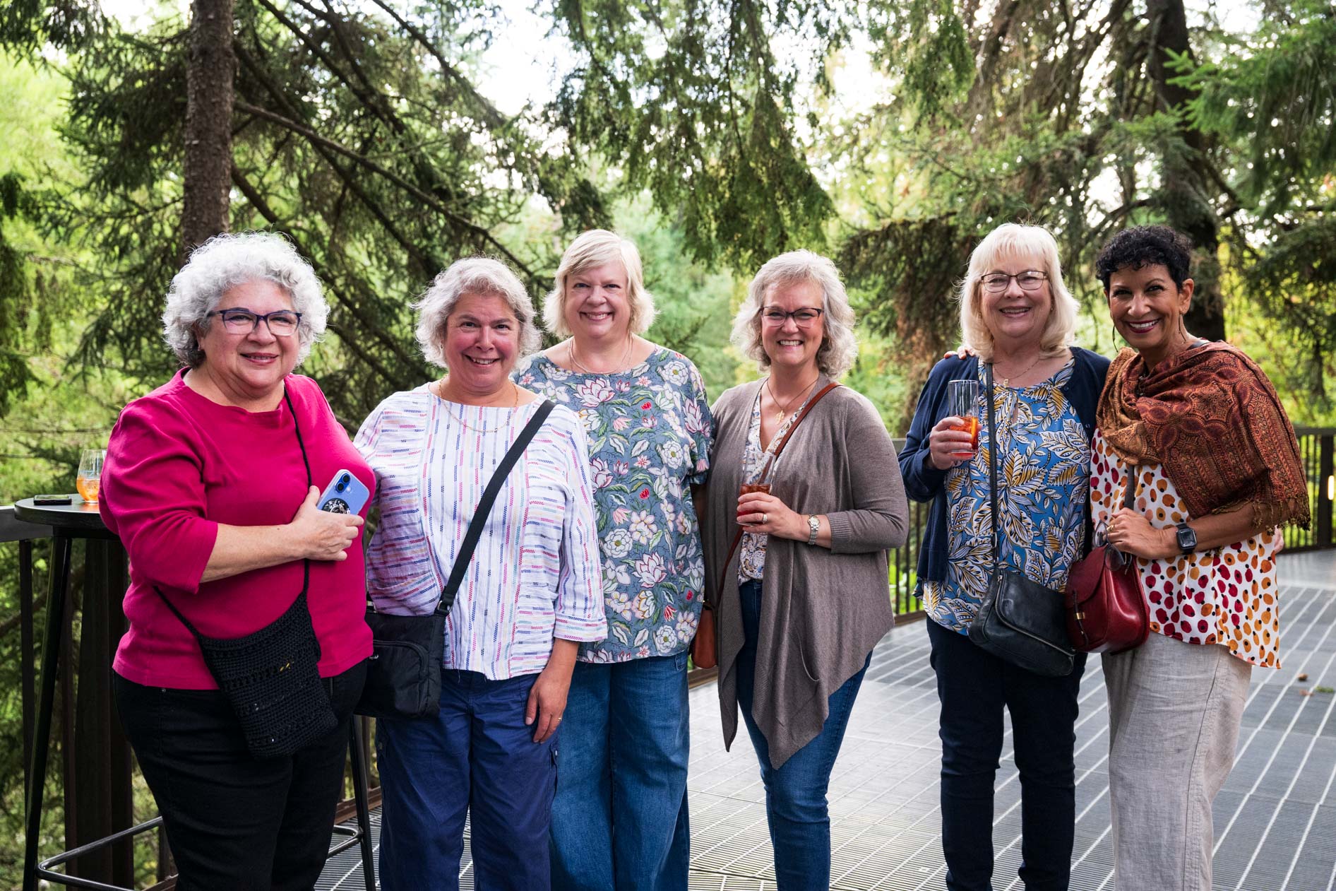  A joyful group of six women poses for a photo on an elevated deck, surrounded by lush trees. The friends are smiling and sharing a moment of genuine connection at the Feast in the Garden event in SE Portland.