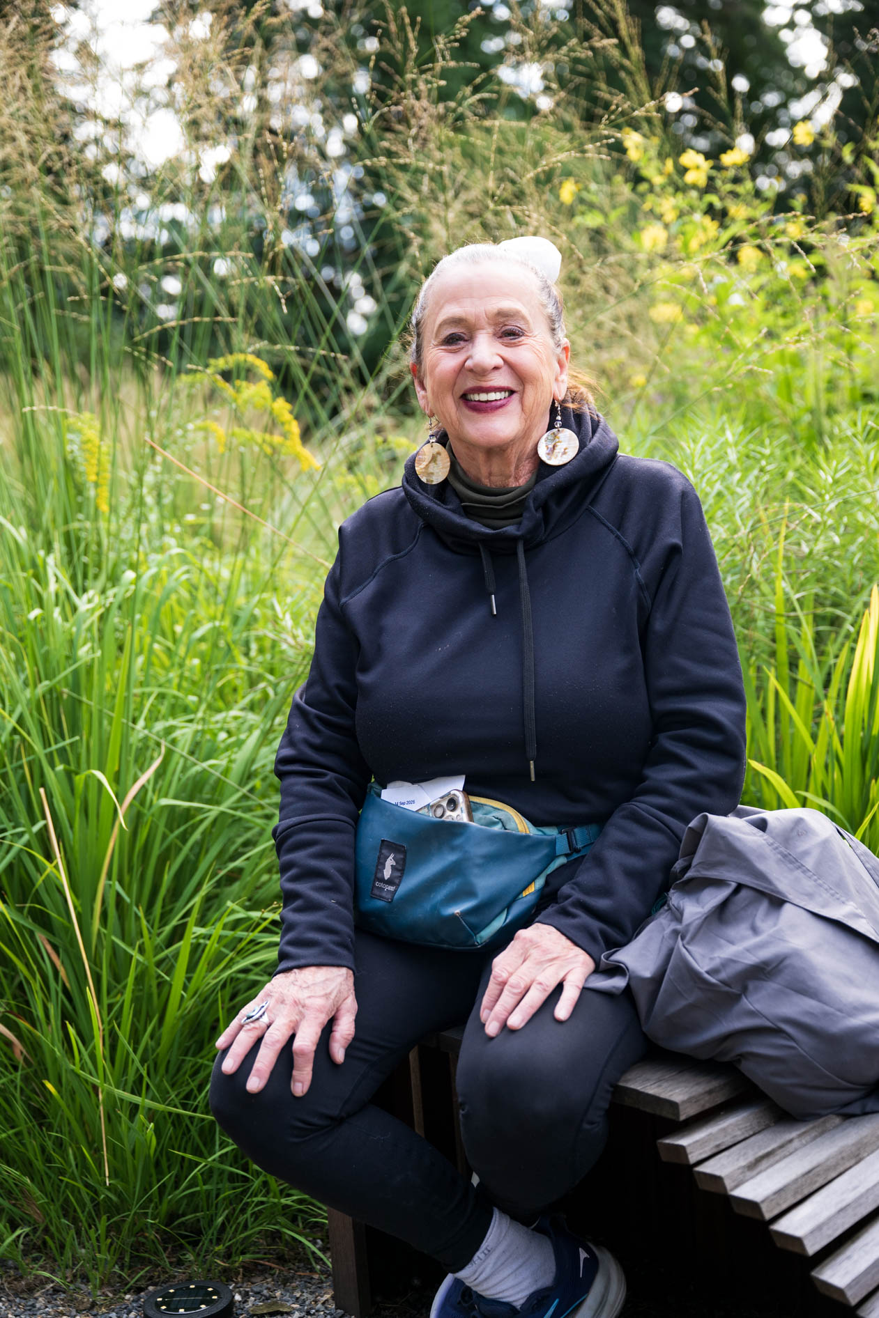  A joyful older woman with a vibrant smile sits on a bench, posing for a photo. She is wearing a black hoodie and a teal fanny pack. The woman's genuine happiness radiates from her face, capturing a serene moment at the Feast in the Garden event in PDX.
