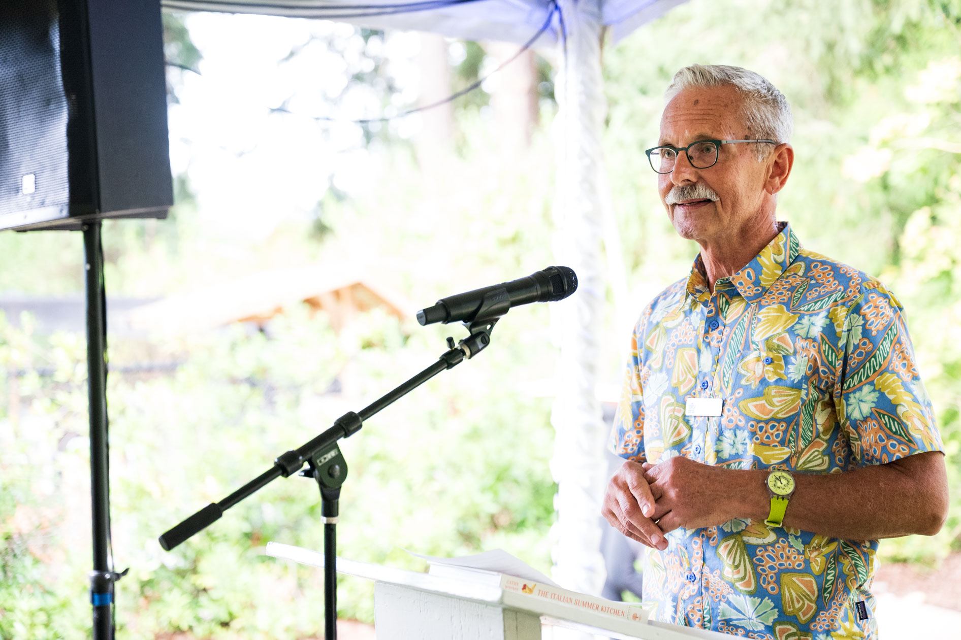 A man with gray hair and a mustache smiles while speaking at a podium with a microphone. He is wearing a vibrant floral shirt, captivating the audience during a speech at the Feast in the Garden event in SE Portland.