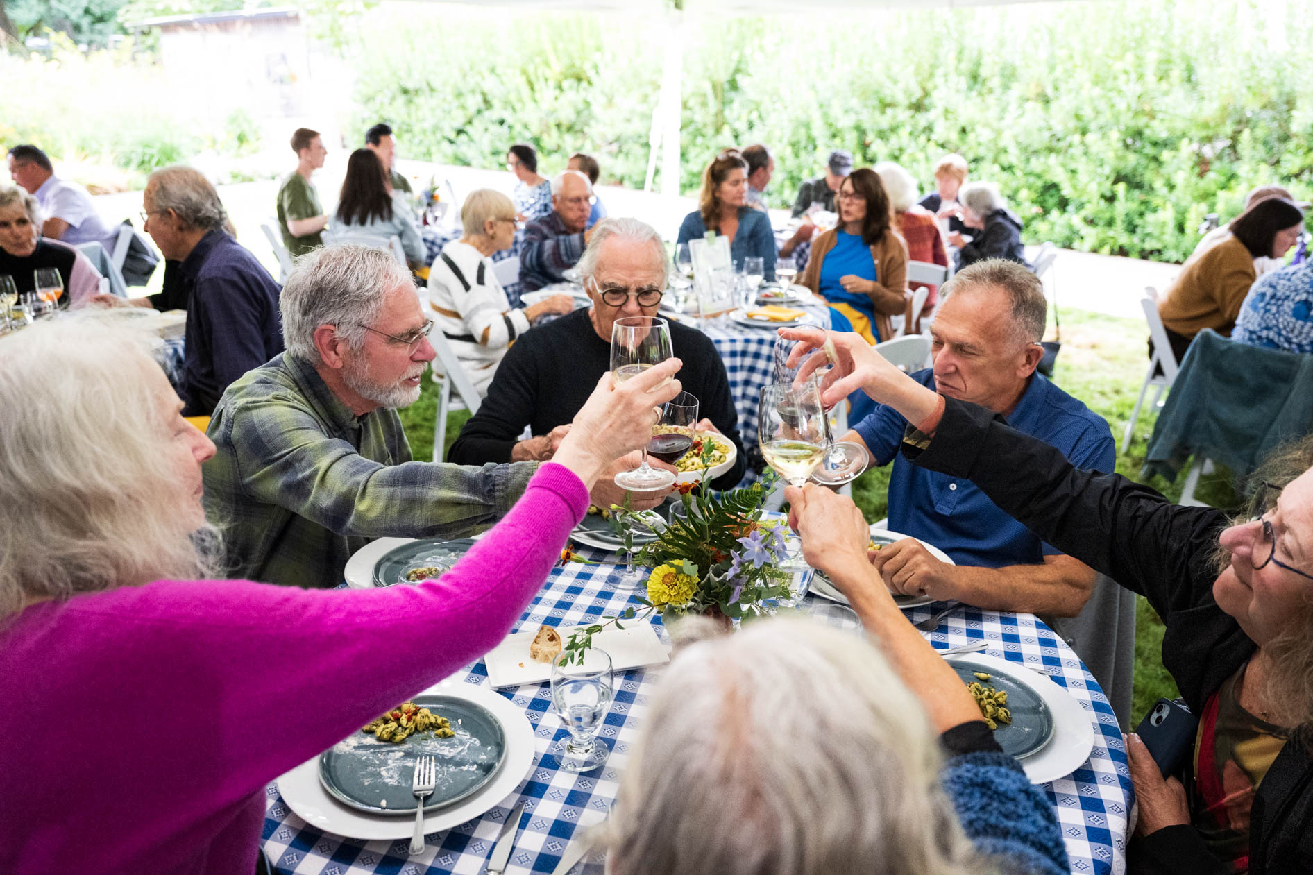 A lively group of guests at a round dining table covered with a blue and white checkered tablecloth. They are toasting with wine glasses, sharing a moment of collective celebration at the Feast in the Garden event in SE Portland.