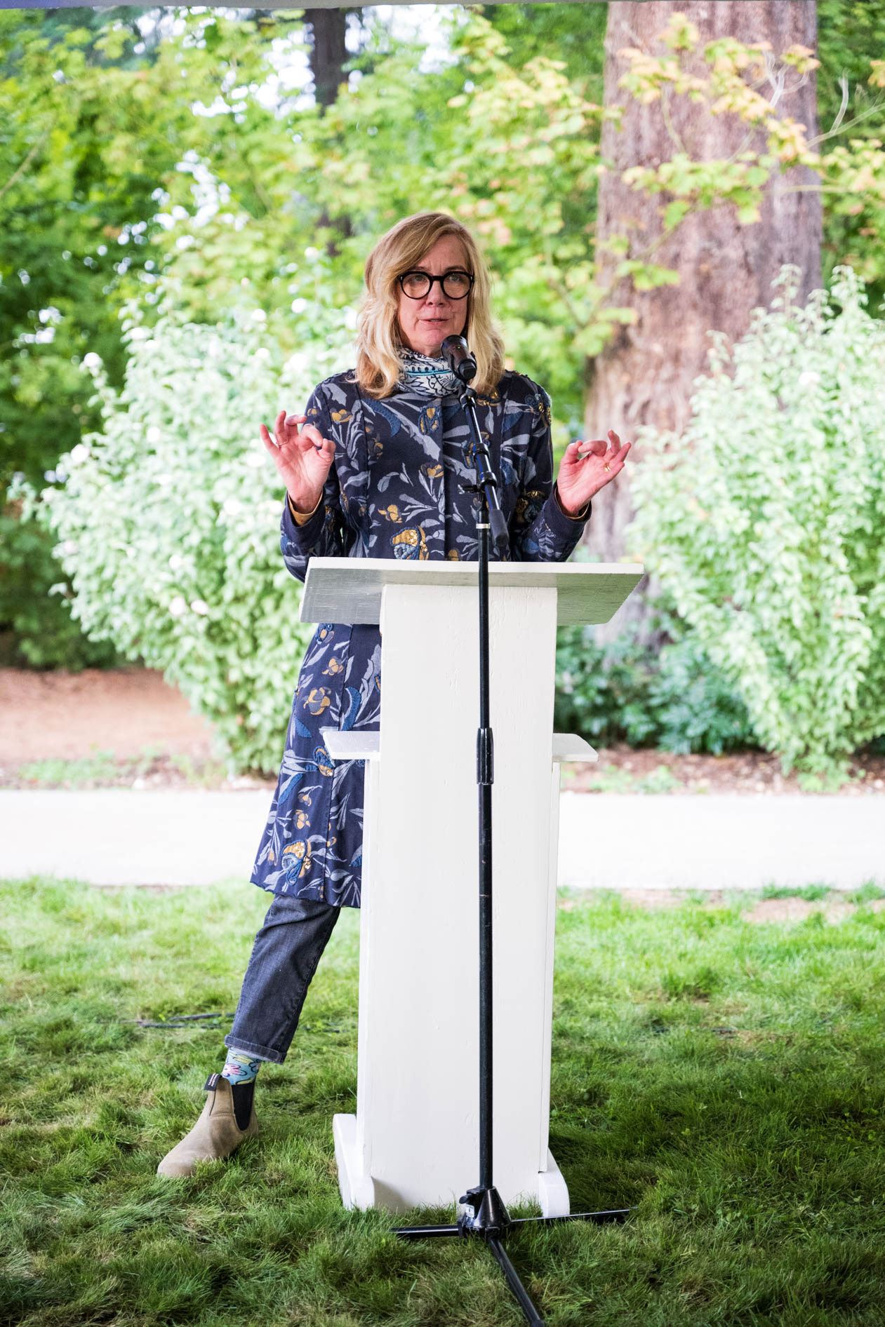A woman with glasses and blonde hair speaks passionately at a white podium, gesturing with her hands. She is wearing a floral coat and standing on a grassy lawn, captivating the audience during a speech at the Feast in the Garden event.