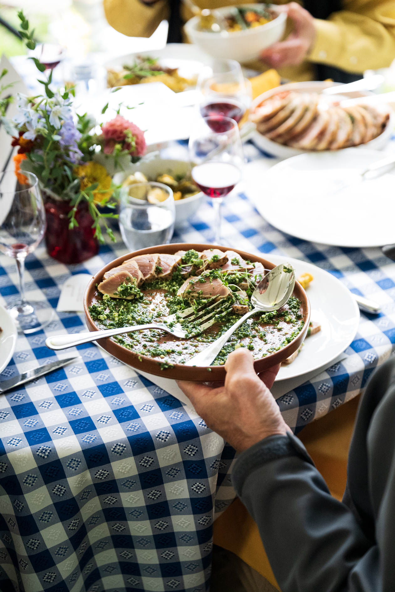 A guest's hands hold a plate with a meal of sliced pork loin and green chimichurri sauce. The plate is over a table with a blue and white checkered tablecloth, capturing the food and intimate dining atmosphere at the Feast in the Garden event in SE PDX.